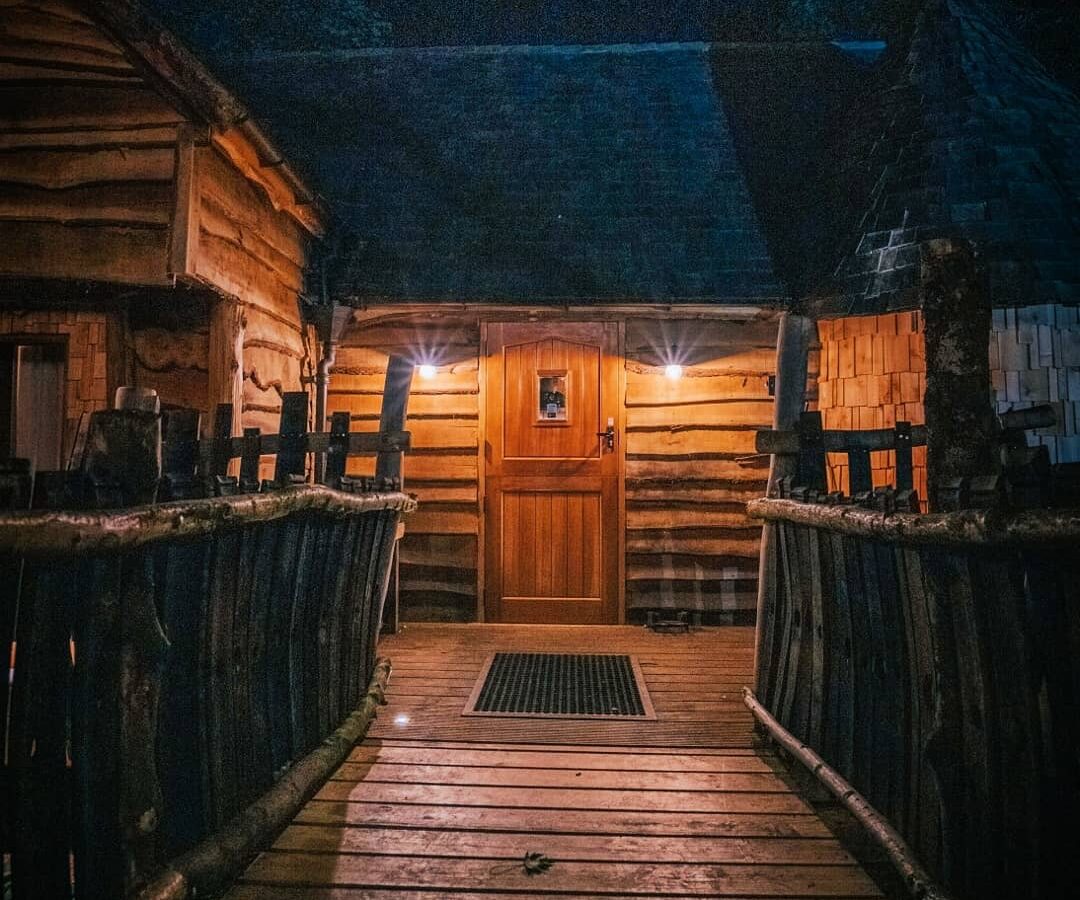 A wooden cabin at night, illuminated by soft lights above the door, reminiscent of Baxby Manor. The cabin is accessed by a rustic walkway made of logs and wooden planks, leading to a closed wooden door. The surroundings appear dark, suggesting a forest or secluded area near Rufus's Roost Treehouse.