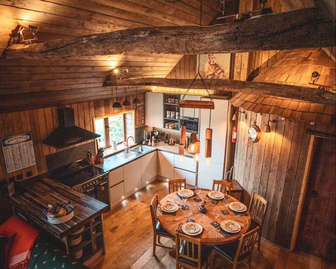 A cozy wooden cabin interior at Baxby Manor features a kitchen area with white cabinets and black appliances, a round dining table set with plates and chairs, and a rustic living space with colorful cushions. Warm lighting and exposed wooden beams create a welcoming atmosphere reminiscent of Rufus's Roost Treehouse.