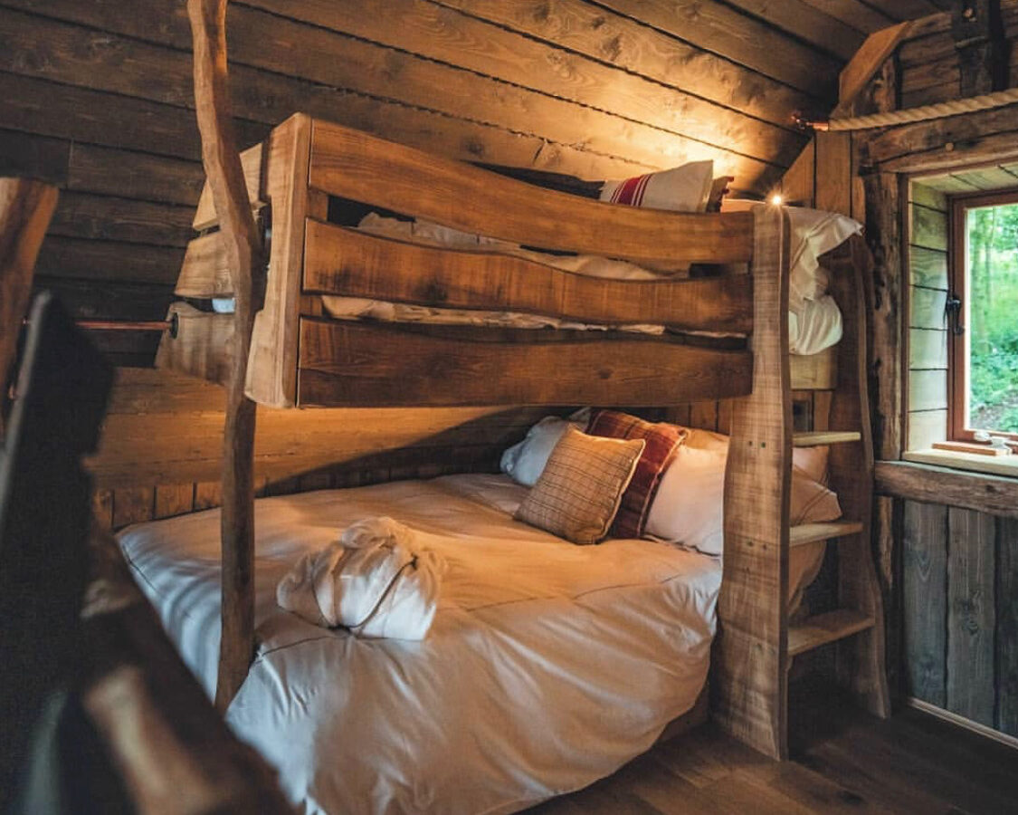 Rustic wooden bunk bed in a cozy cabin room at Baxby Manor. The lower bed is made up with white sheets and pillows, while the upper bunk has a white pillow visible. A small window with a view of greenery lets natural light in. The walls and ceiling are made of wooden planks.