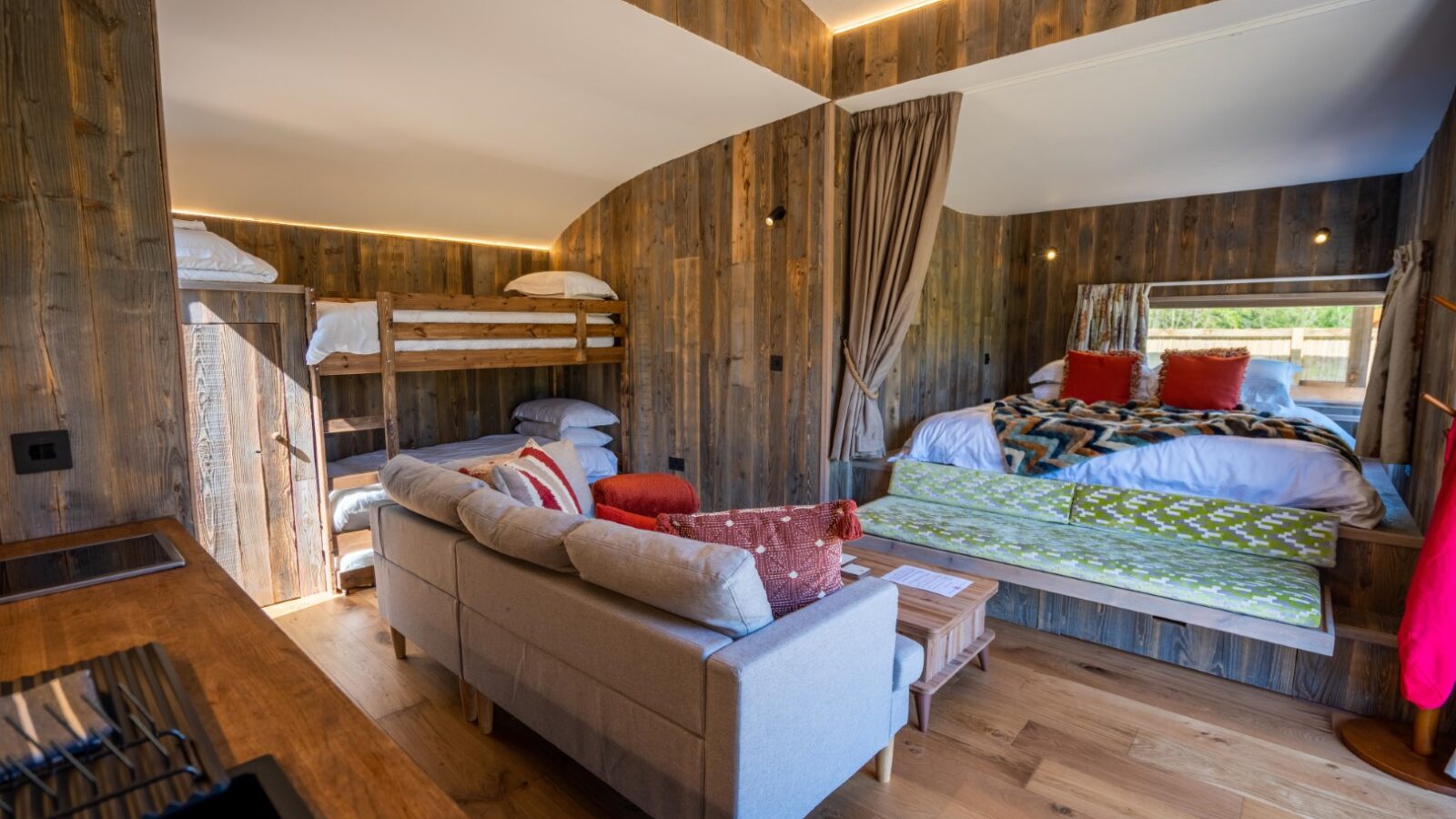 A cozy cabin interior with wooden walls and floors. It features a bunk bed with white linens on the left, a gray sofa with red and orange cushions in the center, and a double bed with colorful blankets on the right. A small window in the corner provides natural light, reminiscent of Baxby Manor's charm.