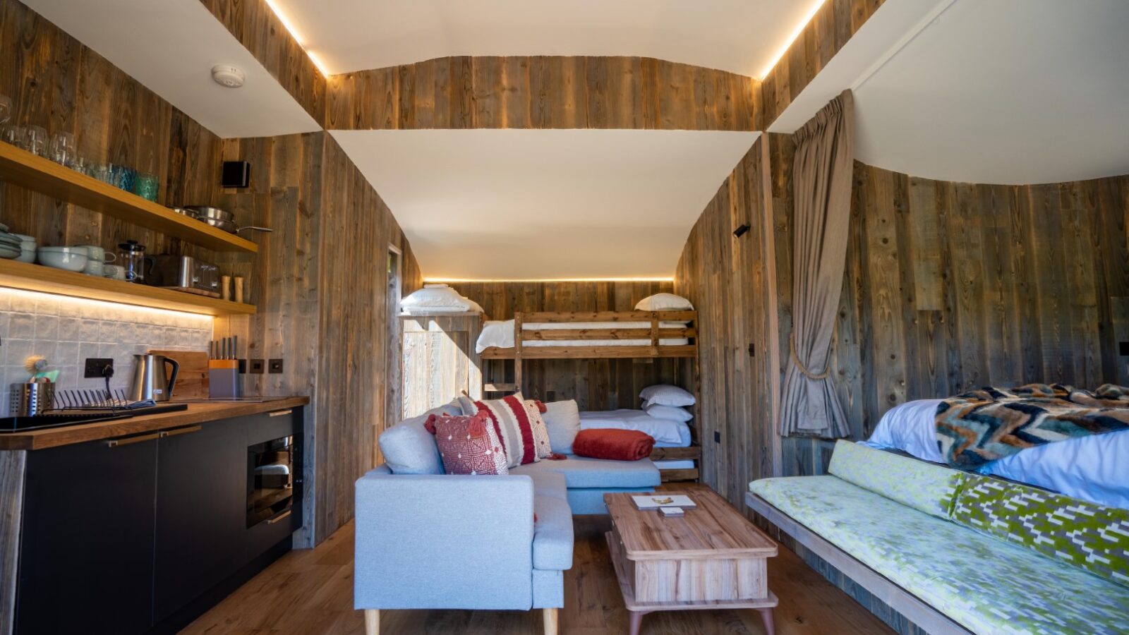 A cozy cabin interior at Baxby Manor featuring wooden walls, a small kitchenette with a black countertop, a light grey sofa adorned with red and white pillows, a wooden coffee table, bunk beds in the corner with white bedding, and a larger bed with a colorful quilt.