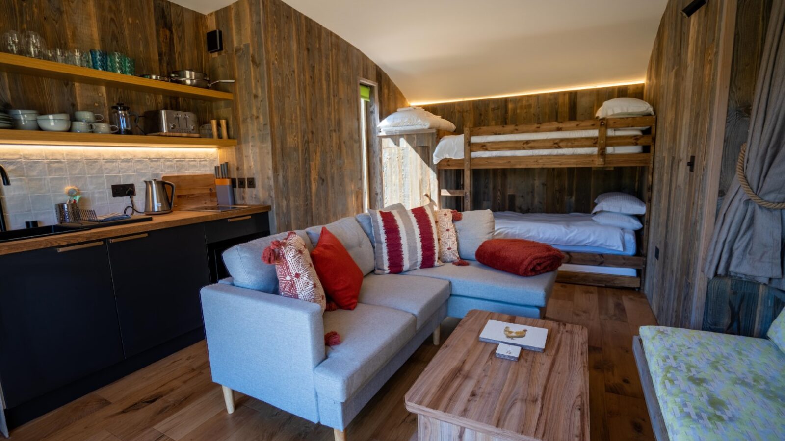 Experience the enchanting charm of Baxby Manor's cozy wooden cabin interior, featuring a kitchen with shelves of dishes, a kettle, and a sink on the left. A Kabinas light gray sectional sofa with red and white pillows and a wooden coffee table are in the center. Bunk beds with white bedding are in the background.