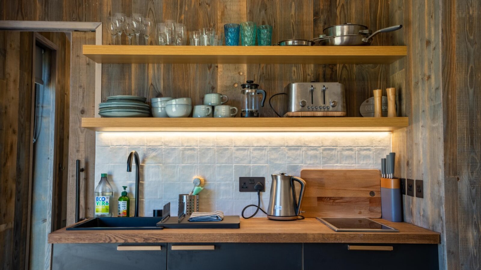 A modern kitchen with rustic wooden walls, reminiscent of Baxby Manor, features open shelves holding glasses, plates, and cookware. The countertop has a sink, a cutting board, a kettle, and various kitchen utensils. There's under-shelf lighting and electrical outlets on the wall.