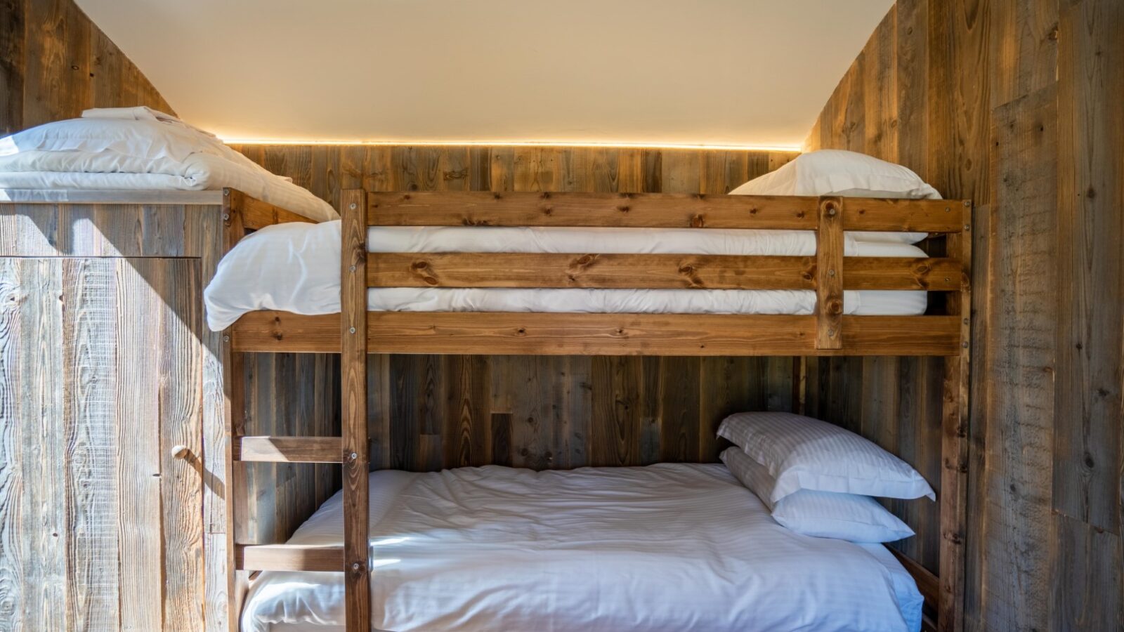 A cozy bedroom at Baxby Manor features a wooden bunk bed with a double mattress on the bottom and a single mattress on top, both neatly made with white bedding. The walls are clad in rustic wooden planks, and soft lighting illuminates the Kabinas room from above.