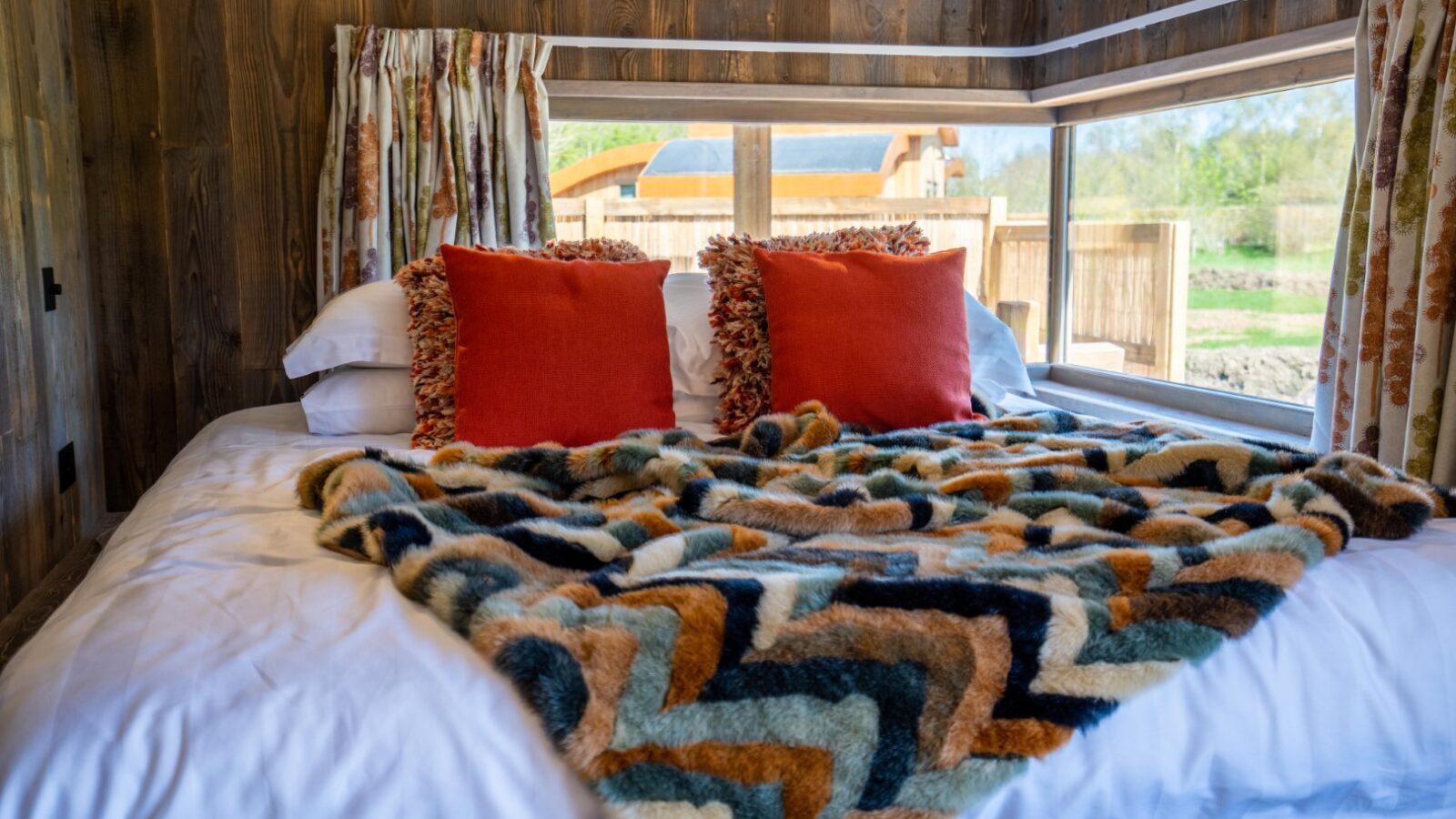 A cozy bedroom at Baxby Manor with a wooden interior features a large bed adorned with a colorful, zigzag patterned blanket and bright orange throw pillows. The bed is positioned near a corner window, offering a scenic view of the outdoors with greenery and another building visible.