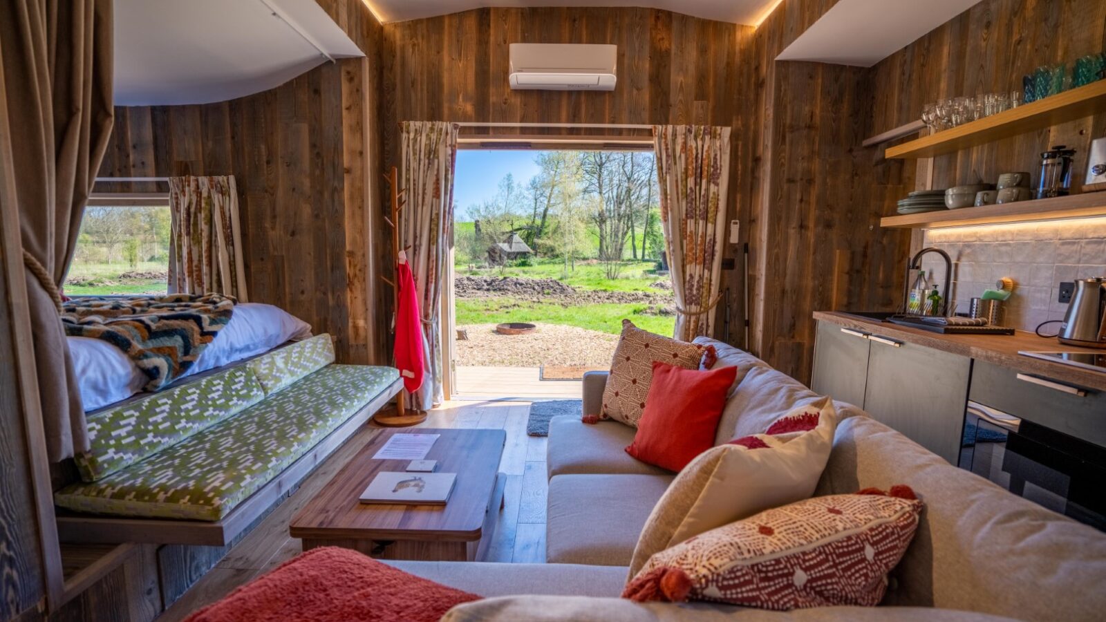 A cozy, rustic tiny home interior at Kabinas features a sofa with red and patterned cushions, a small kitchen area, and a raised bed with a colorful blanket. Large windows and an open door frame a natural outdoor view, letting in abundant daylight. Wood accents cover the room.