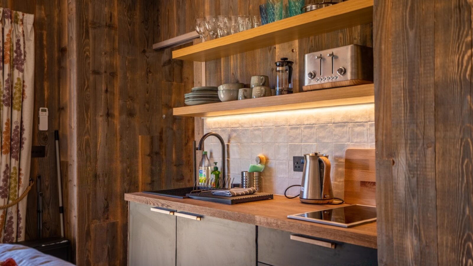 A cozy kitchen at Baxby Manor with wooden walls and shelves. The countertop features a sink with a faucet, a kettle, and a tablet. Shelves hold glassware, plates, and a coffee maker. A wooden backsplash with under-shelf lighting adds warmth to the space. A curtain is partially visible.