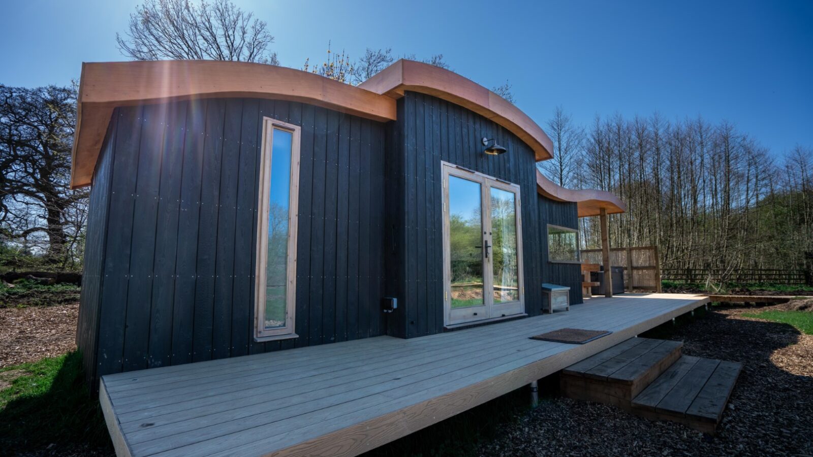 A modern tiny house with wooden exterior and large windows stands on raised wooden decking at Baxby Manor. The roof features a unique curved design. In the background, a clear blue sky, trees, and sunlight streaming from the top left corner enhance the natural beauty of Kabinas. Wood chip paths surround the area.