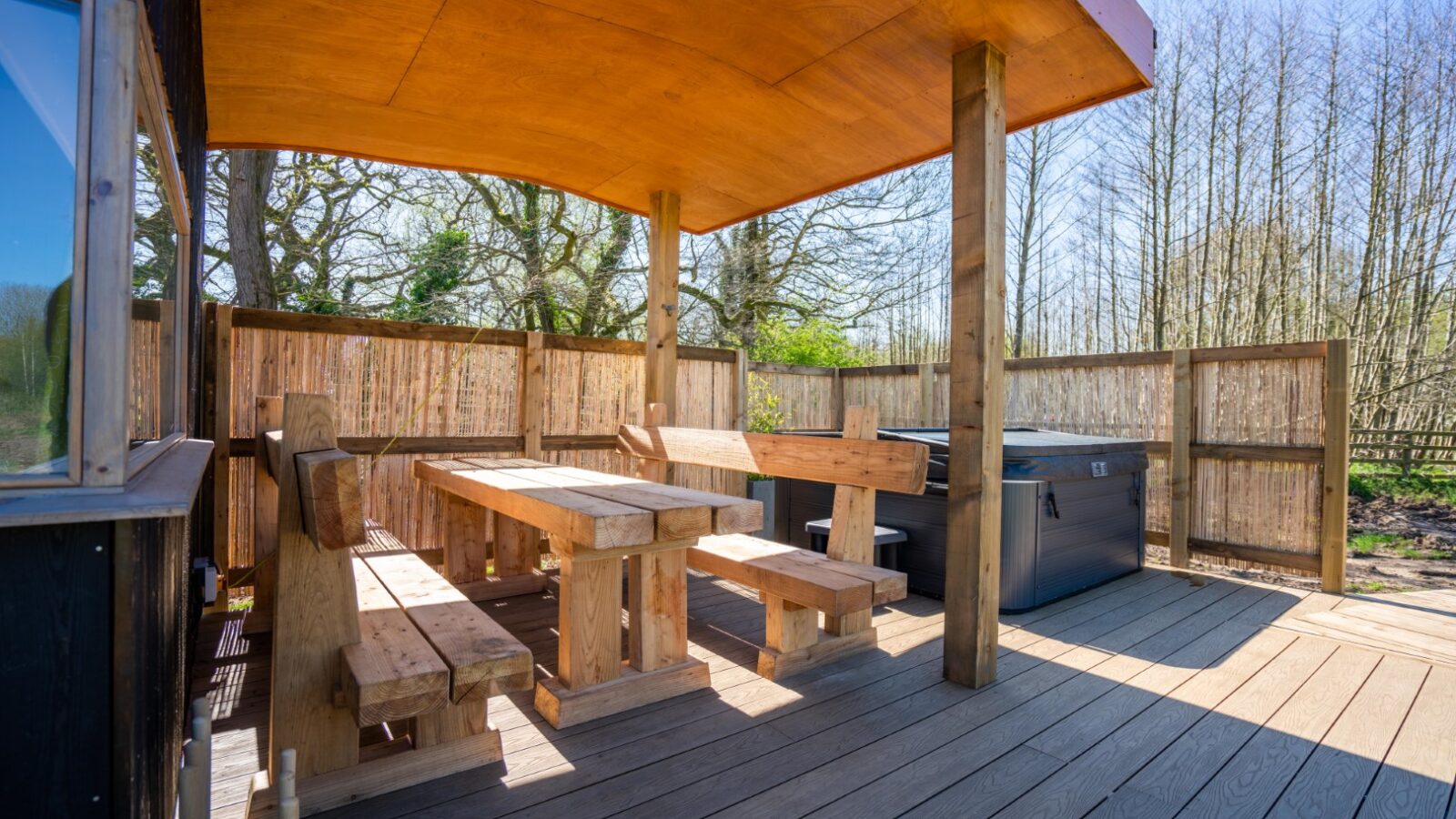 A spacious outdoor deck at Baxby Manor features wooden furniture including a picnic table and benches. Covered by a wooden roof and enclosed by a slatted fence, the deck also boasts a hot tub on one side. With trees in the background, it creates a serene, nature-filled setting perfect for Kabinas guests.