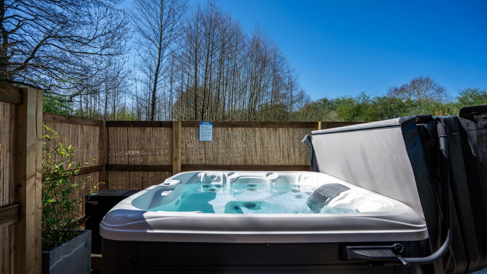 A hot tub filled with water is surrounded by a wooden fence and trees at Baxby Manor. The sky is clear and blue, with the partially open cover hanging to the side. Green foliage and Kabinas are visible in the background, providing a serene outdoor setting.