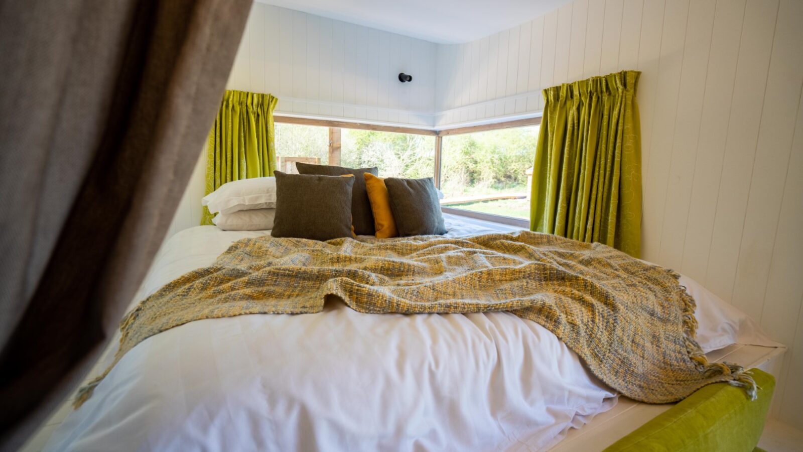 A cozy, well-lit bedroom at Baxby Manor with a cushioned bed adorned with white sheets, a textured blanket, and multiple throw pillows. The room features large windows with green curtains, allowing natural light to illuminate the space and provide a view of the outdoors.