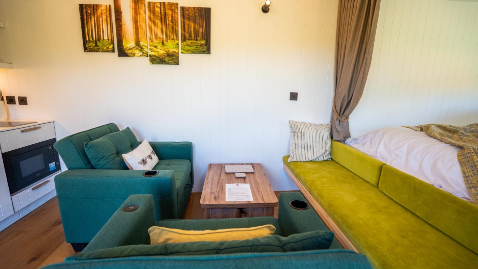 A cozy living space at Baxby Manor features a teal armchair and loveseat, a wooden coffee table, and a green cushioned bench. A wall art piece depicting a sunlit forest hangs above the loveseat, and a small portion of a bed and curtain are visible on the right.