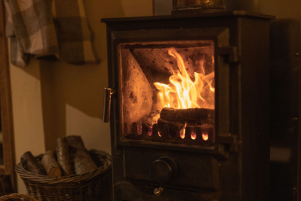 A cozy wood-burning stove with a fire ablaze inside, emitting a warm glow. Logs are stored in a woven basket to the left, and a checkered cloth hangs on the wall. This scene at Cobnut Cabin evokes warmth and comfort, suggesting a rustic, homey atmosphere perfect for Ritty Retreats' cabin rentals.