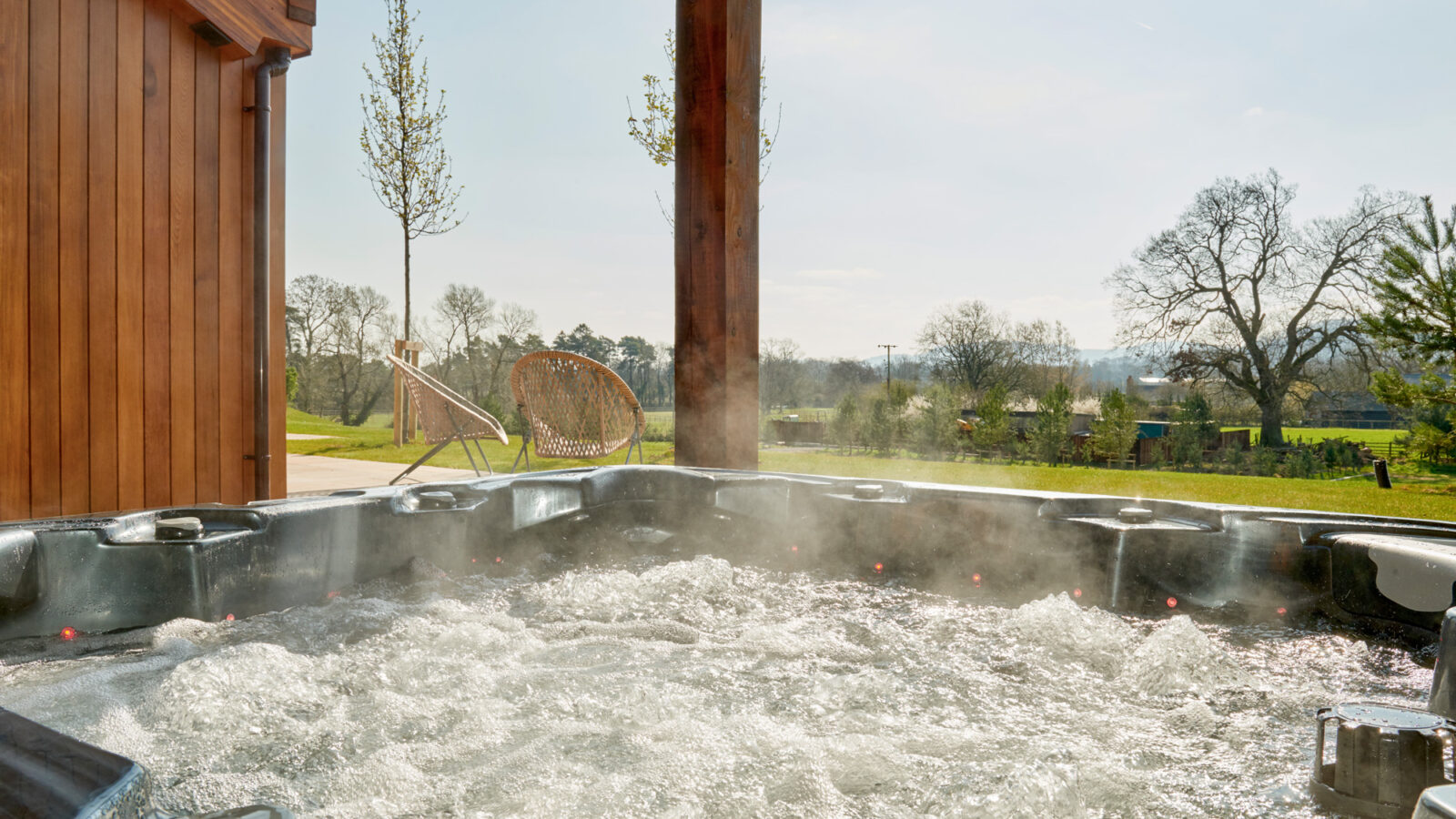 A steaming hot tub is seen under a wooden cover with a picturesque background of trees, parkland, and clear skies. A cozy hanging chair is positioned near the hot tub, adding to the serene outdoor setting often found at Parkland Lodges.