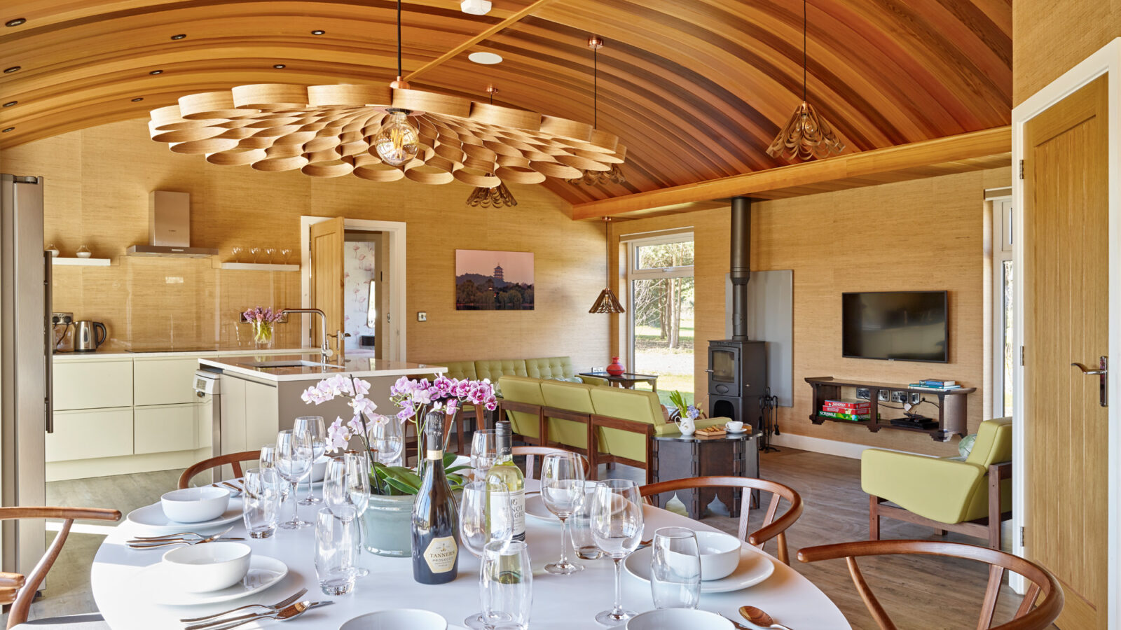 A modern dining area in a parkland-inspired lodge with a wooden curved ceiling, featuring a white circular table set with plates, bowls, cutlery, and wine glasses. There’s wine and flowers on the table. The background includes a kitchen, a wood stove, a wall-mounted TV, and lime-green seating.