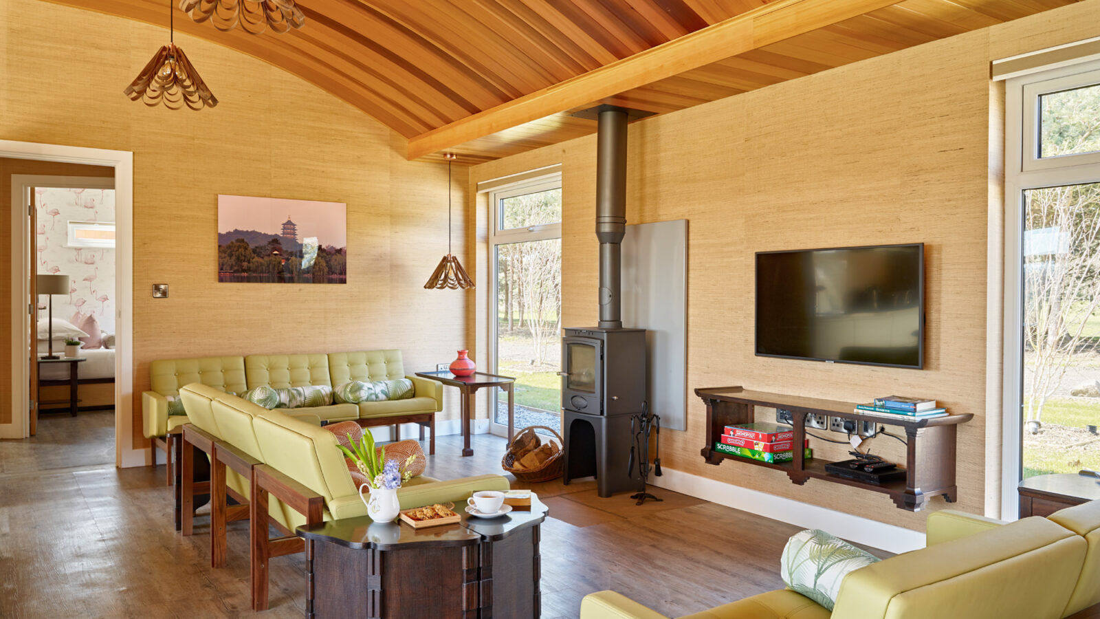 A cozy living room in this charming lodge features a wooden ceiling and light wooden floor. The space includes green cushioned seating, a black wood-burning stove, a wall-mounted TV, and a bookshelf. A door on the left leads to a bedroom. Large windows bring in ample natural light from the surrounding parkland.