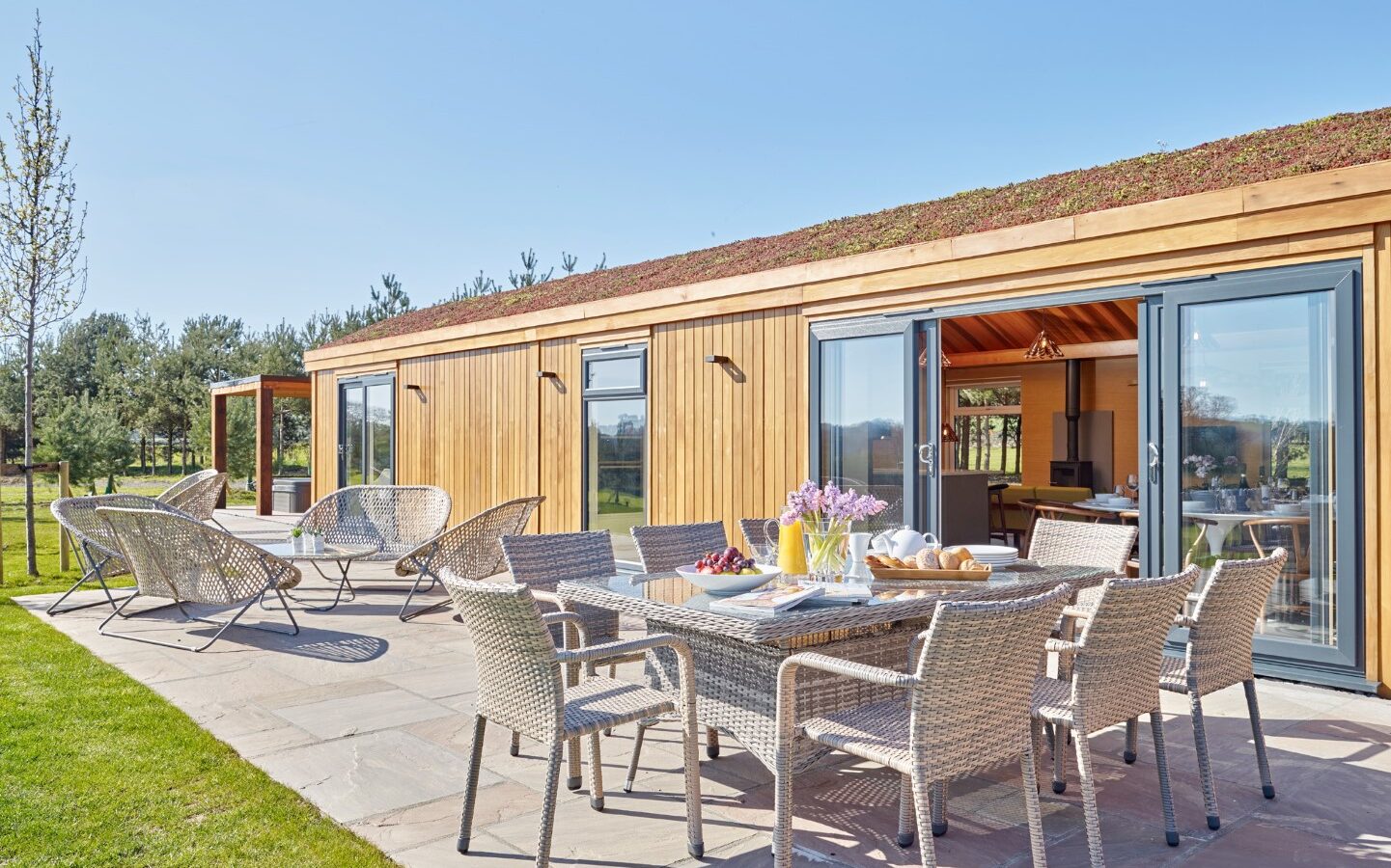 A modern, single-story wooden house with large glass sliding doors stands gracefully among the parkland. The outdoor space features a paved patio area furnished with wicker chairs and tables, set for a meal. The surrounding area has lush greenery and a bright, clear sky overhead, making it an ideal lodge setting.