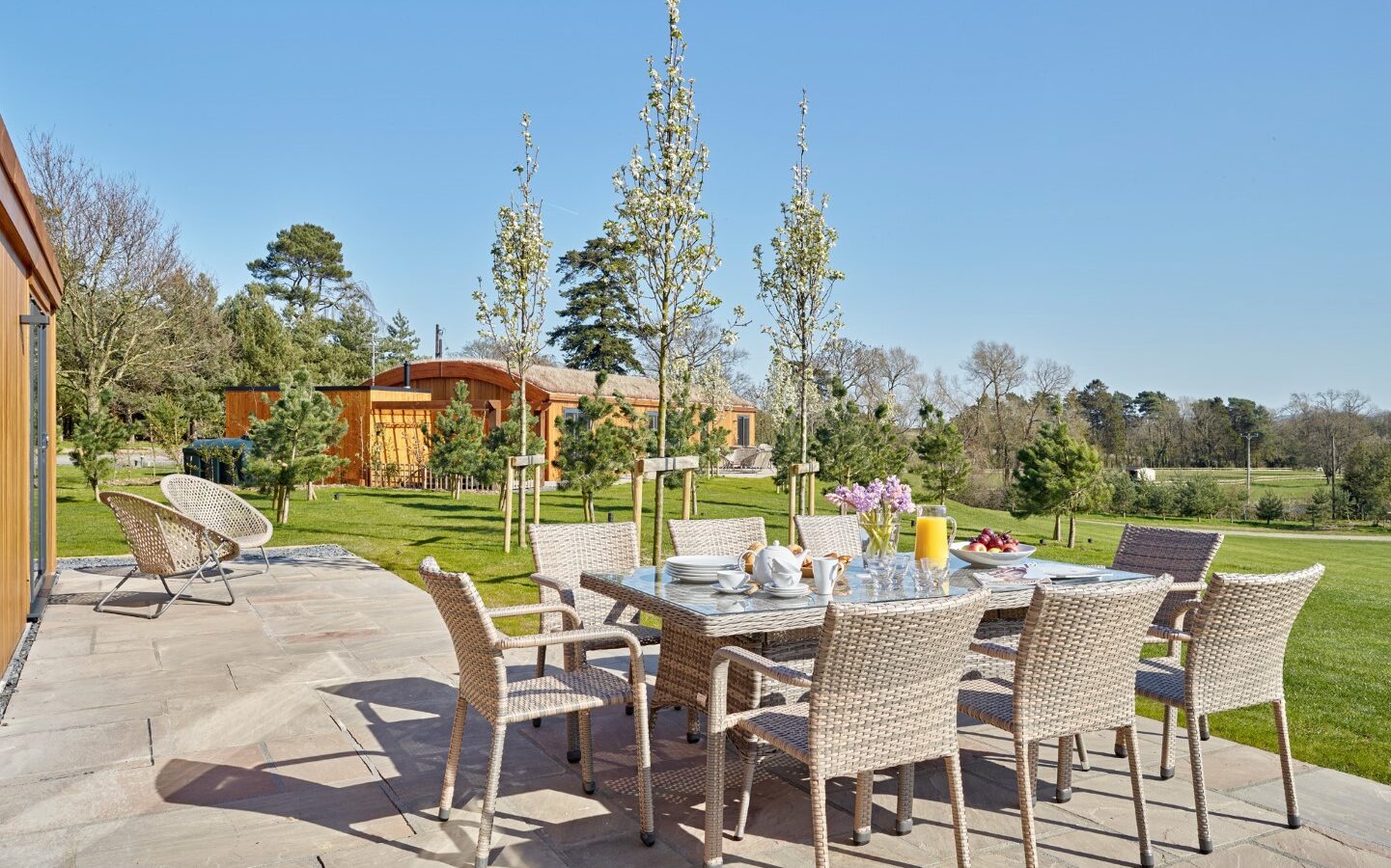 Outdoor patio featuring a dining table set for a meal with wicker chairs arranged around it. Located on the stone-paved terrace of Parkland Lodges, the scene is bathed in bright sunlight with three young trees in the background, under a clear blue sky and surrounded by lush greenery.