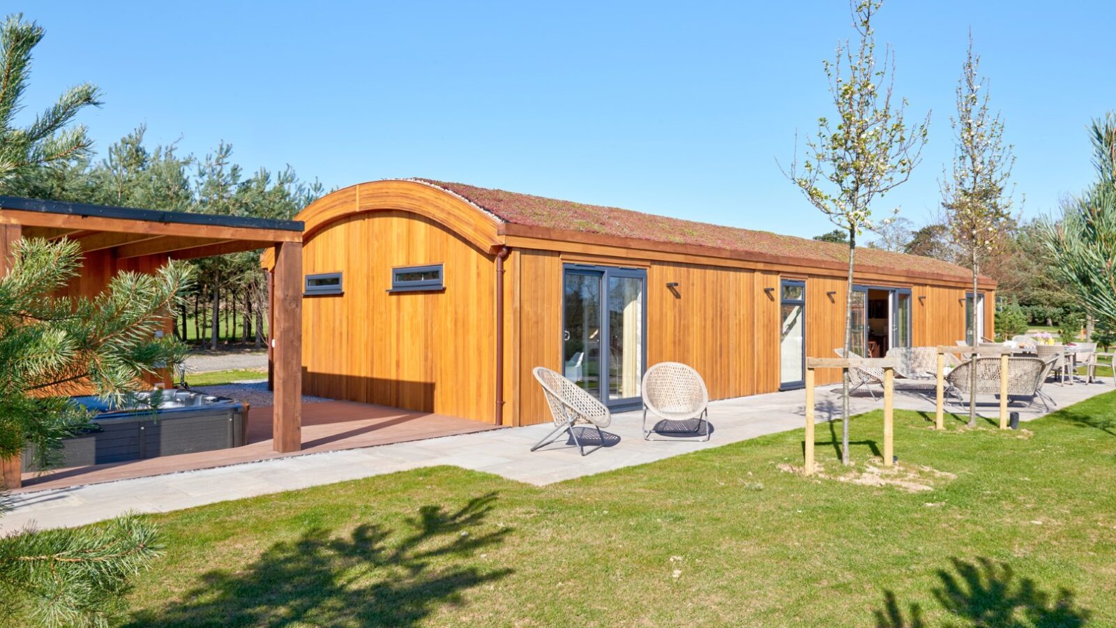 A modern, single-story wooden cabin with a curved roof sits under a clear blue sky in a serene parkland. The patio features wicker lounge chairs and a hot tub. The surrounding area includes lush green grass, young trees, and scattered outdoor furniture, making it one of the coziest lodges around.