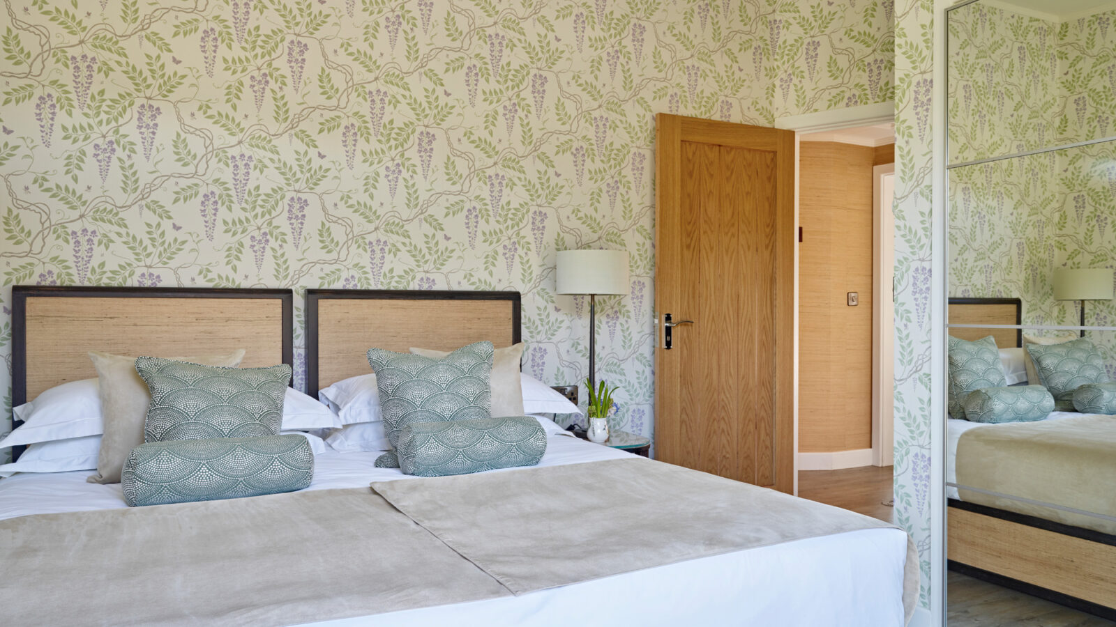 A neatly made bed with two light green decorative pillows and light gray bedcovers is positioned in a cozy bedroom adorned with floral wallpaper at Parkland Lodges. A wooden door is slightly open, and a mirrored wardrobe reflects the room. A floor lamp stands by the bedside.