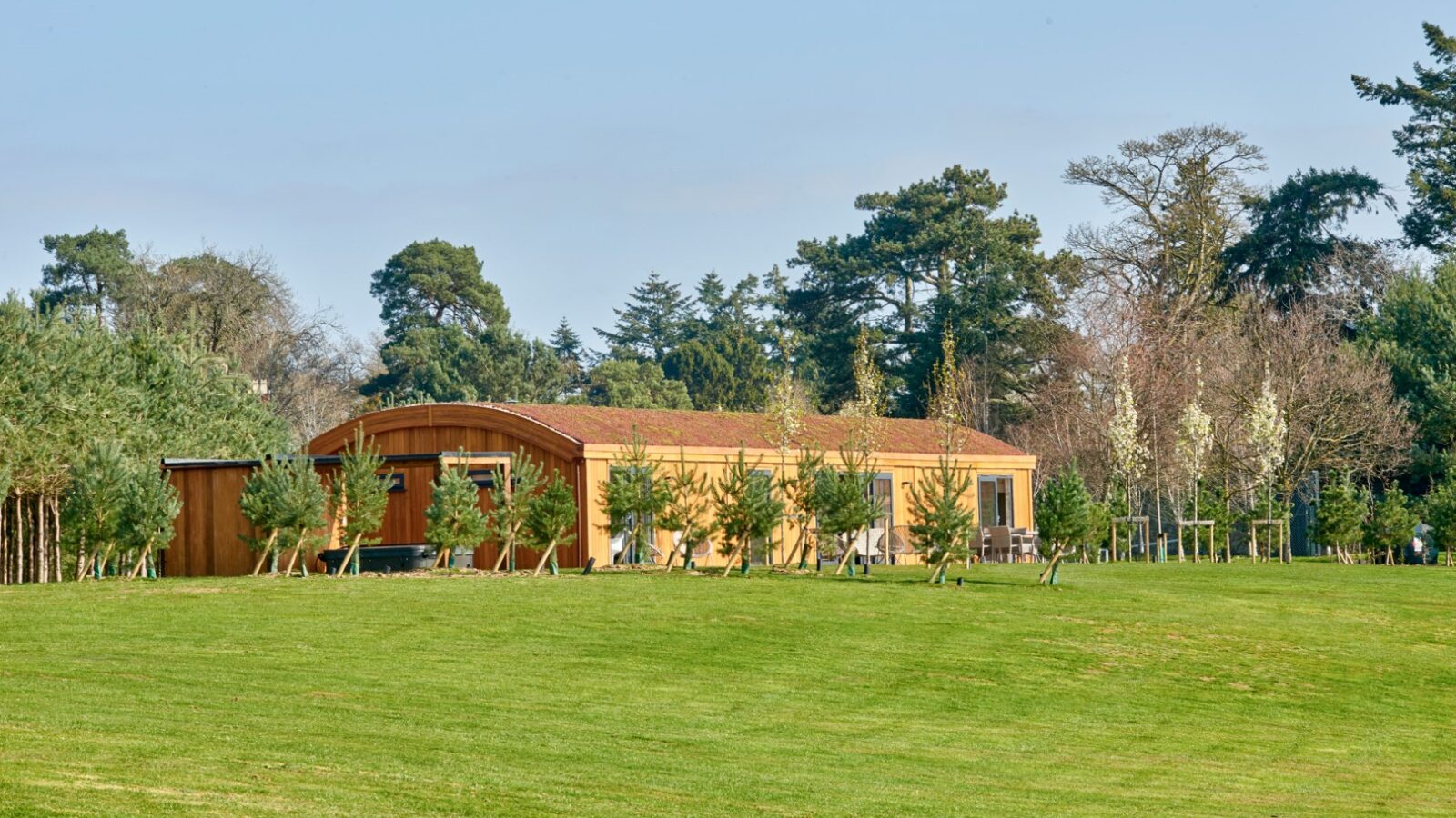 A modern, single-story building with a curved roof, featuring large windows, is situated in a landscaped grassy area. Surrounding the building are small trees and shrubs, with taller trees and a clear blue sky in the background, making it one of the charming Parkland Lodges.