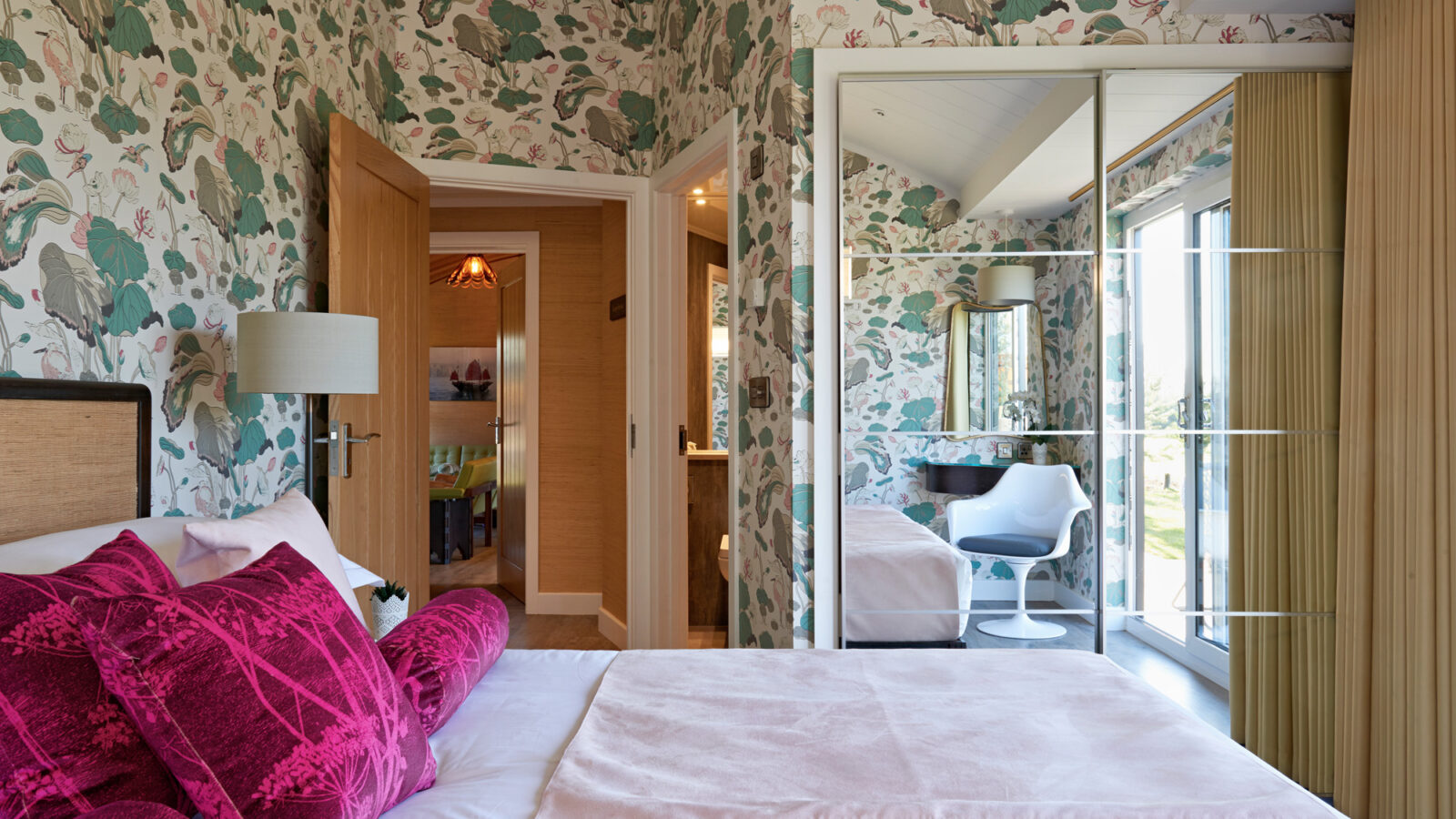 A bedroom with floral wallpaper, a white bed with pink pillows, and a mirrored closet on the right. A door leads to an en-suite bathroom. There is a white chair and a window overlooking the serene parkland outside, filling the room with natural light, perfect for lodges.