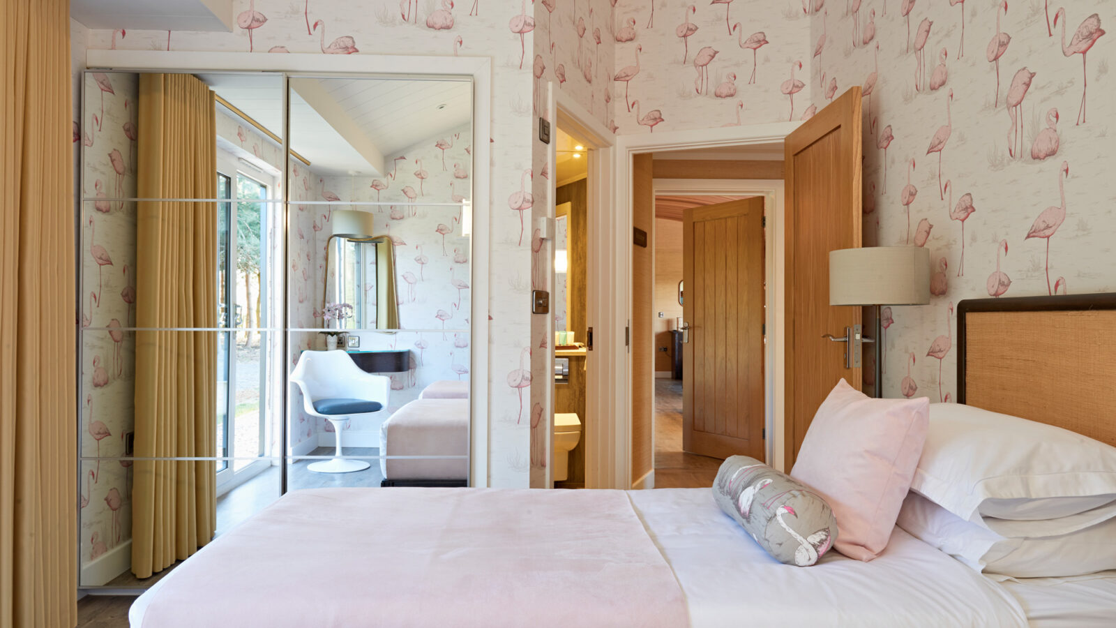 A cozy bedroom at Parkland Lodges with flamingo-themed wallpaper features two neatly made twin beds with pink accents. The room has a wooden door, a mirrored wardrobe, and a vanity with a white chair by the window. Warm lighting and neutral tones create a welcoming atmosphere.