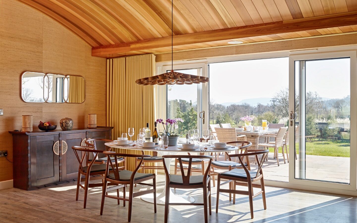 A bright, modern dining room with a wooden arched ceiling graces the interior of Parkland Lodges. A large round table set with tableware is surrounded by wooden chairs. Sliding glass doors open to an outdoor patio with a dining set and a scenic view of a green landscape and distant hills.