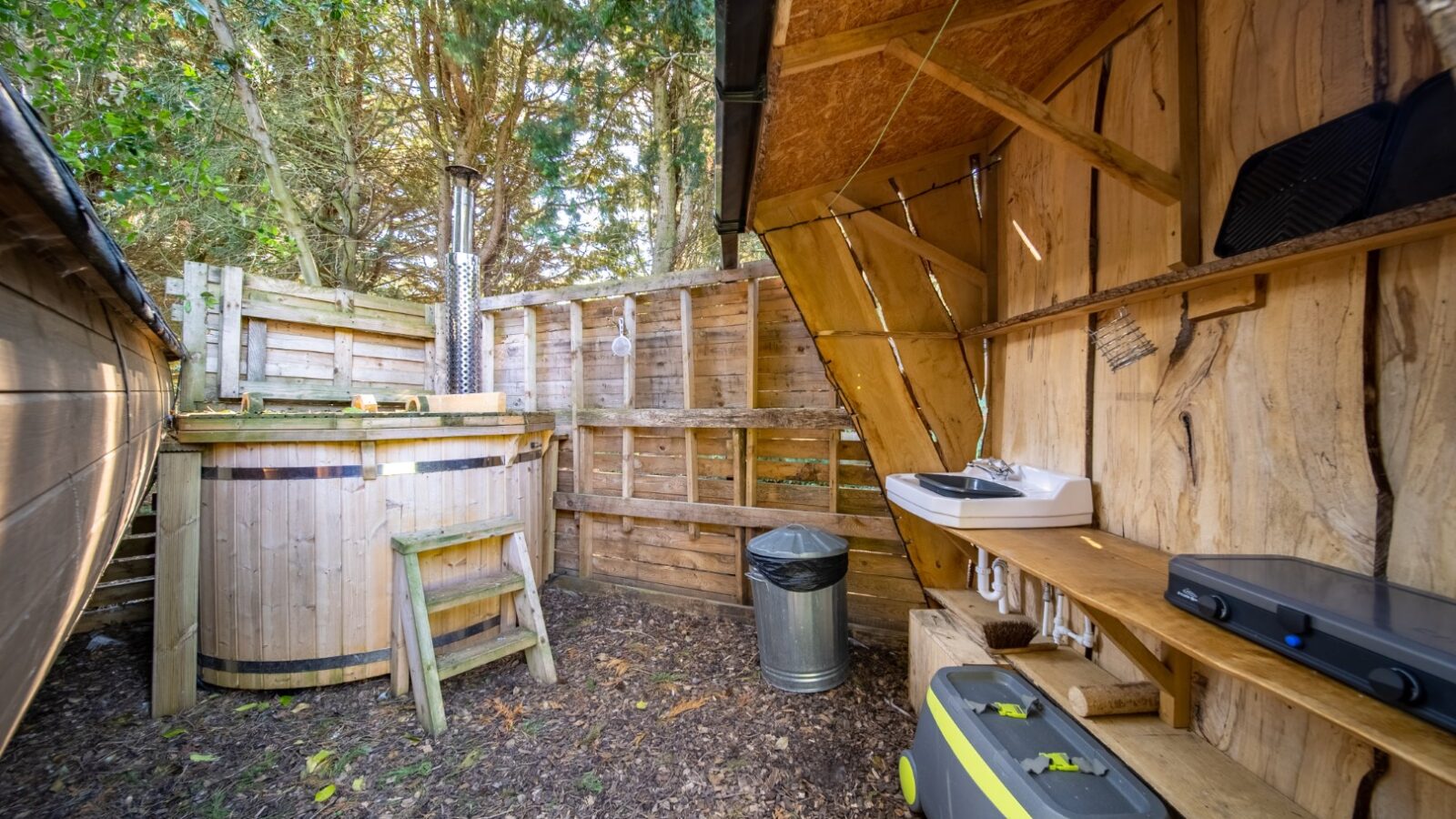 An outdoor wooden hot tub area with steps leading into the tub, perfect for a glamping retreat. A wooden fence surrounds the space, and on the right is a small outdoor sink with a faucet. A small wooden shelf holds some equipment, and below the shelf sits a green and gray cooler.