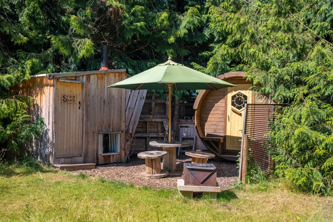 A cozy outdoor seating area surrounded by tall trees at Lahtle Wood features a rustic wooden sauna and a small wooden cabin, perfect for glamping. In the center are wooden tables and stools shaded by a large green umbrella, with a small fire pit nearby. The scene is serene and inviting.