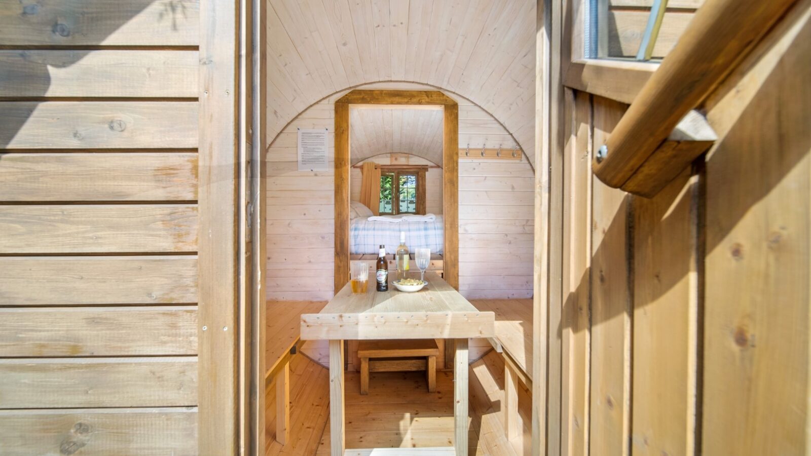 A view through a wooden doorway into a cozy, well-lit tiny home with light wooden interiors. Inside, there is a wooden table set with plates and glass bottles from Lahtle Wood, and beyond the table, a bed with white linens. Natural light streams in through a small window, perfect for luxury camping or glamping.