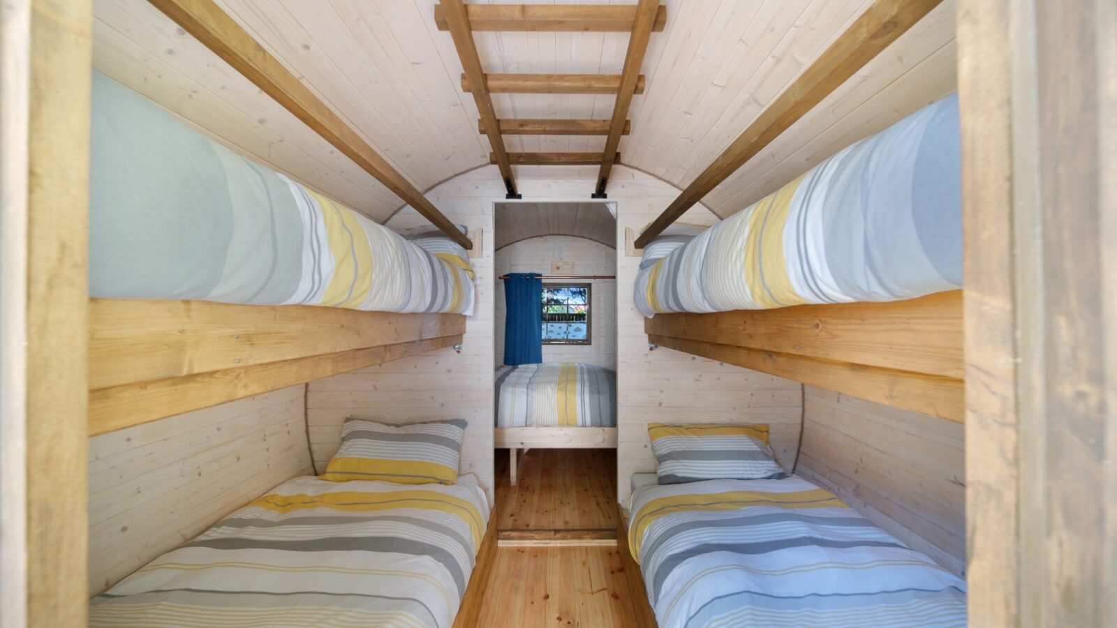 A cozy Lahtle Wood cabin interior with four single bunk beds, two on each side, and a full-sized bed at the back of the room. The walls, ceiling, and floor are made of light wood. The beds are neatly made with striped bedding in shades of blue, white, and yellow. Perfect for your next glamping adventure!