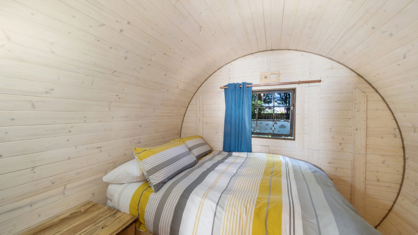 A cozy wooden room with curved walls and a modern, minimalist aesthetic offers a perfect glamping experience. The room features a neatly made bed with striped bedding in white, gray, and yellow. A small window with a blue curtain allows natural light to enter. There's a wood bedside table on the left.