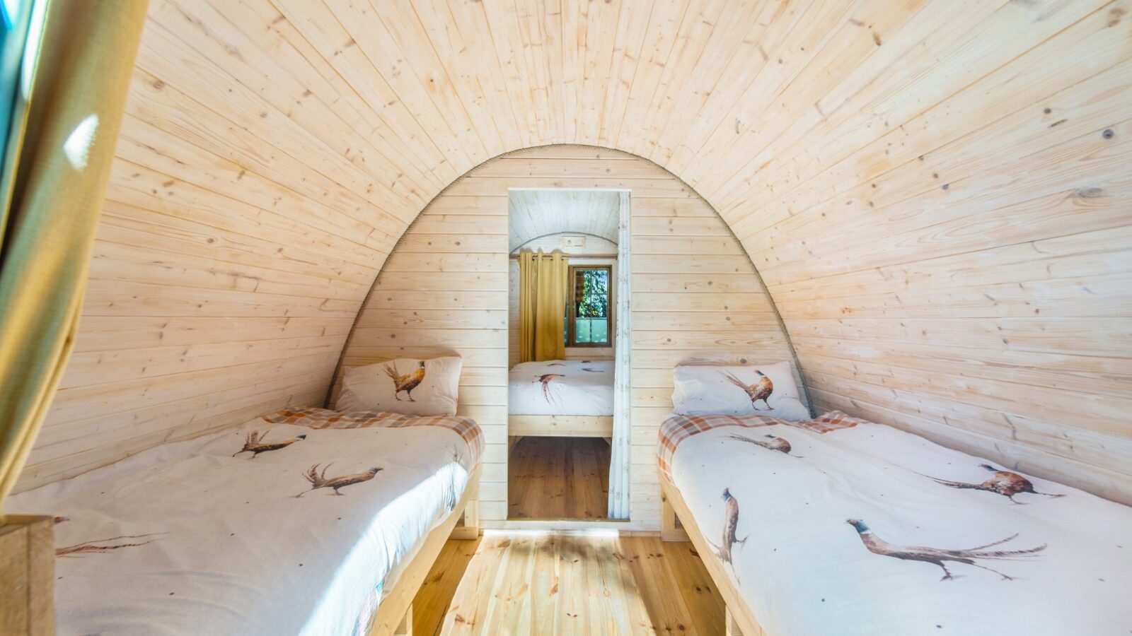 A cozy wooden cabin interior with curved walls, featuring two single beds adorned with bird-themed duvet covers on either side of a central hallway. The Lahtle Wood light finish enhances the bright, inviting atmosphere, perfect for glamping aficionados. A small mirror reflects the corridor.