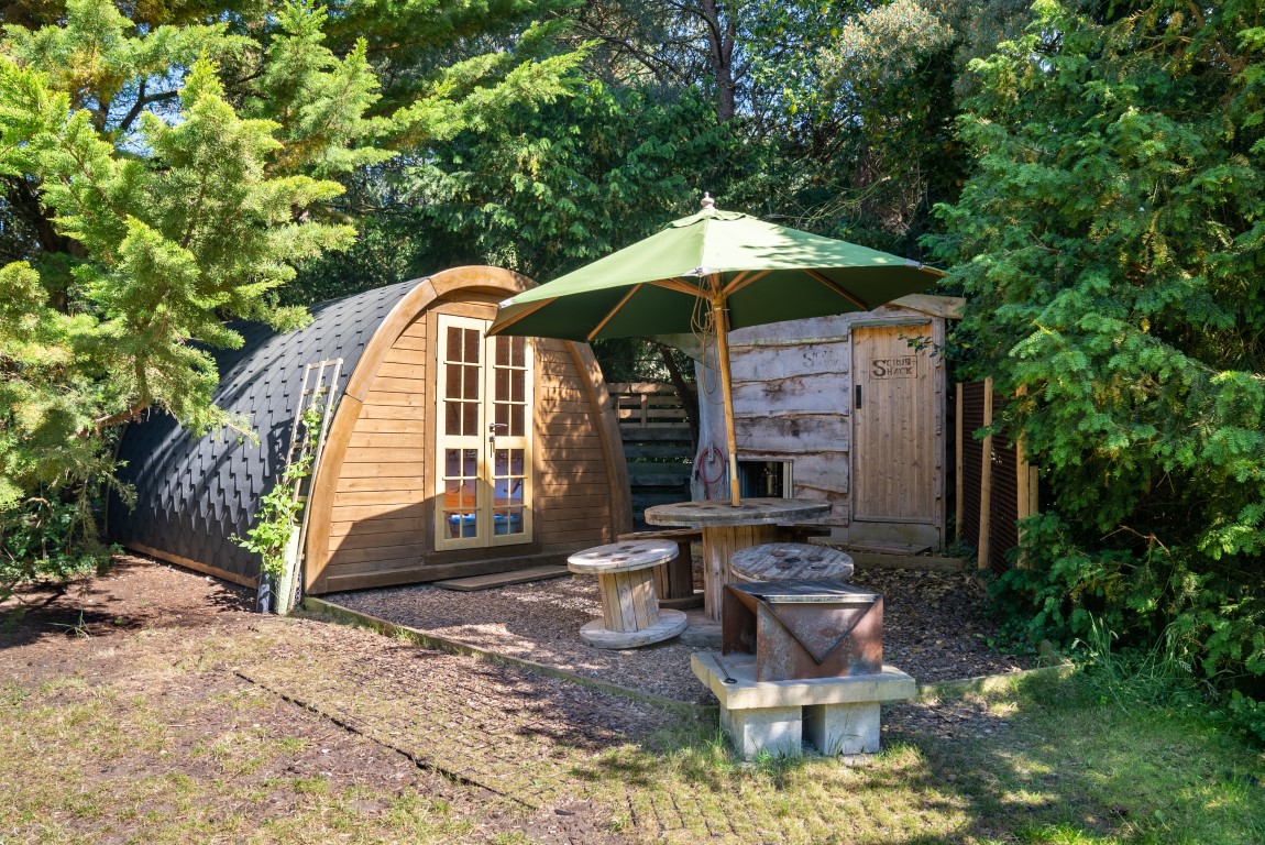 Nestled in Lahtle Wood, this cozy wooden cabin with a curved roof is the perfect glamping retreat. Surrounded by lush greenery, it features a glass door, a small patio with wooden tables and stools, and a large green umbrella for shade. A rustic outdoor shower area adds to the charm on the right.