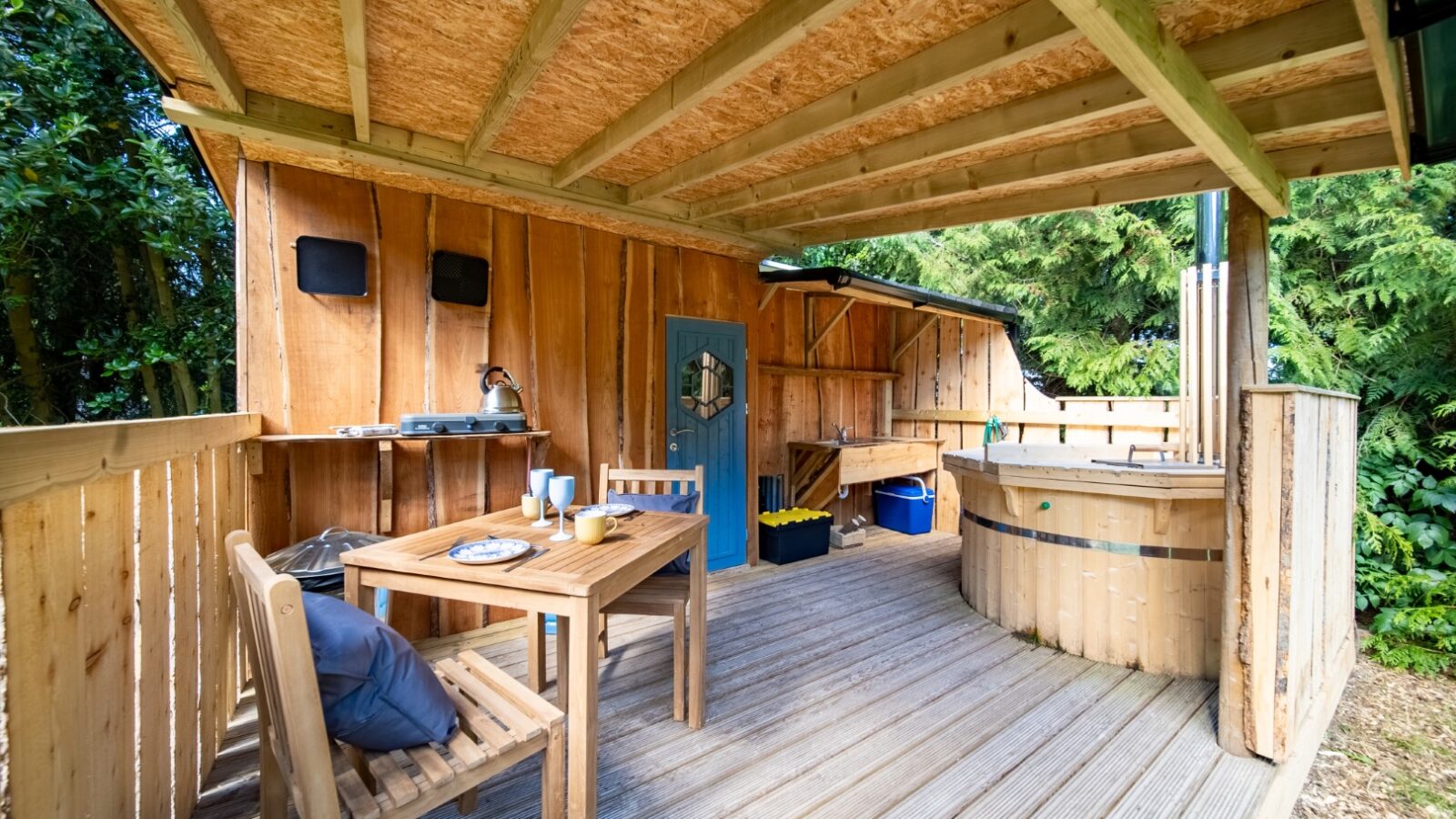 A cozy Lahtle Wood outdoor patio features a small dining area with a table, two chairs, and a hot tub on the right. The space is surrounded by lush greenery. Perfect for glamping, the patio has a wooden roof and includes a workbench with stools and kitchen utensils.