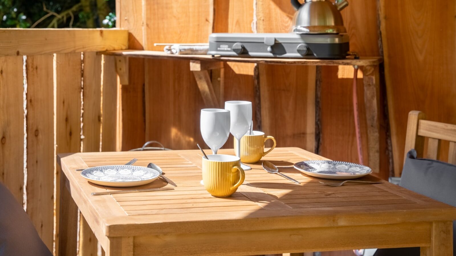 A cozy outdoor wooden dining area at Lahtle Wood's glamping site features a table set for two. The table has two white plates, two yellow mugs, two wine glasses, and cutlery. A silver kettle heats on a portable stove in the background while sunlight filters through the open wooden walls.