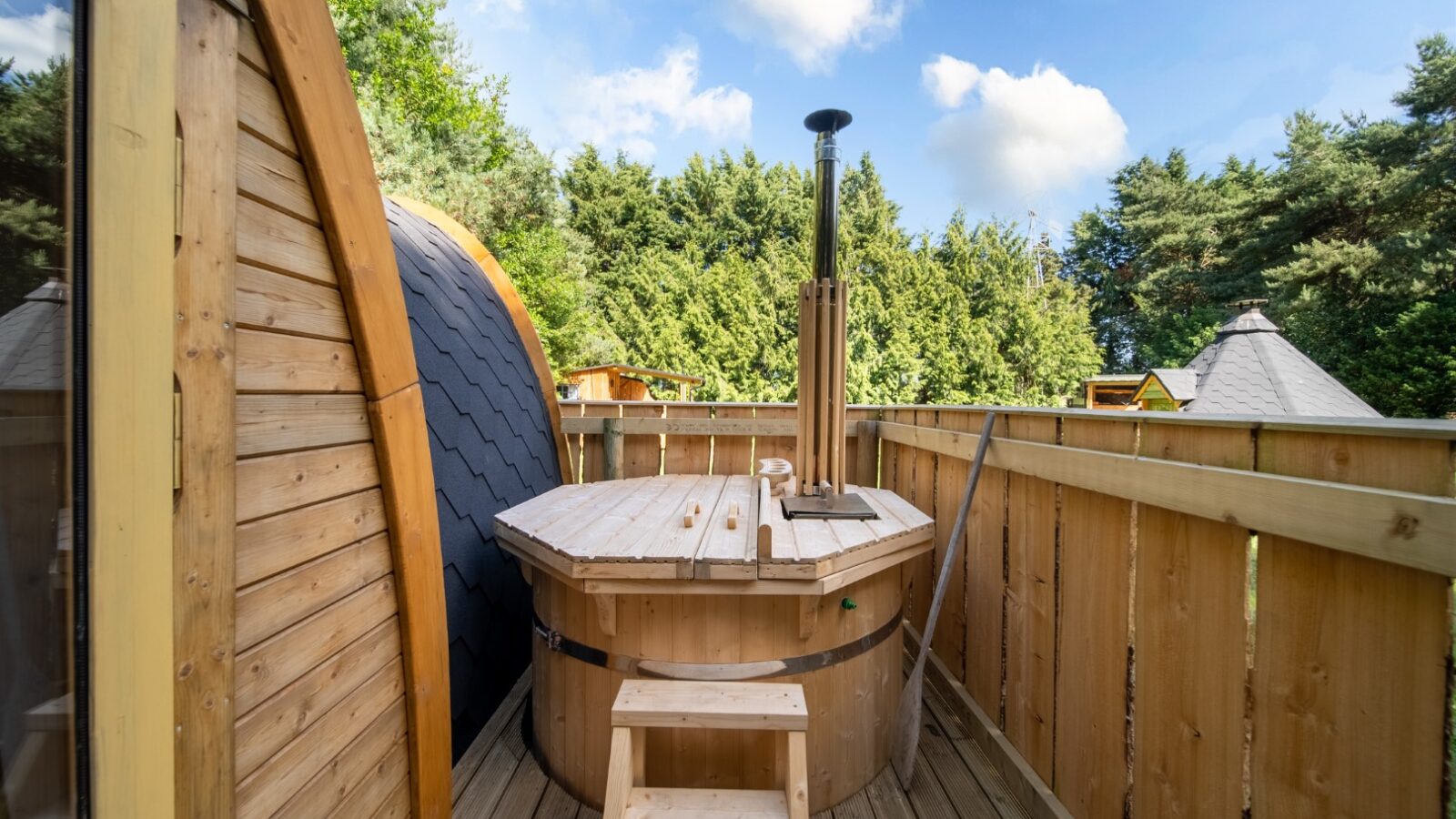 A Lahtle Wood Glamping wooden hot tub with a small chimney sits on a wooden deck. A wooden fence encloses the deck. There is a dome-shaped wooden structure on the left, and trees are visible in the background under a blue sky with scattered clouds.