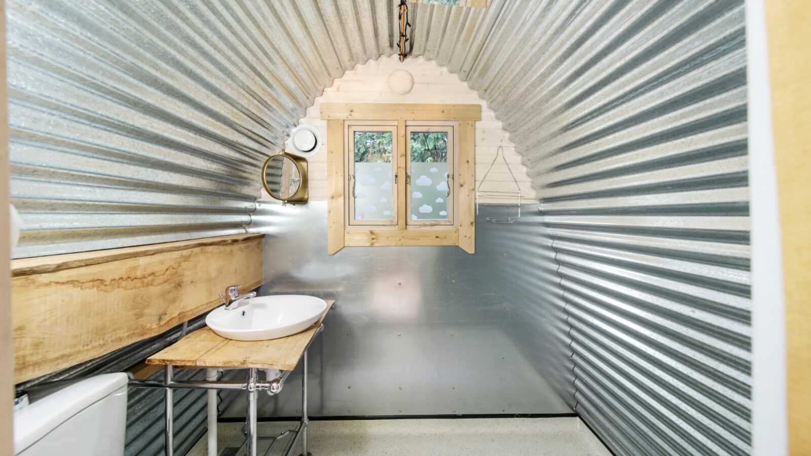 A small, rustic bathroom with corrugated metal walls evokes the charm of wood glamping. A wooden-framed window with frosted glass sits above a wall-mounted sink and mirror. There is a toilet on the left and a wooden countertop on metal legs beside it. The space is lit by a small round light.