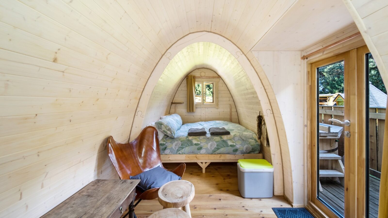 A cozy, wooden, dome-shaped cabin interior featuring a neatly made bed with patterned bedding. The space includes a wooden table, a leather butterfly chair, two small round stools, and a door leading outside with a view of greenery. Perfect for glamping enthusiasts, the room has a warm, inviting atmosphere.