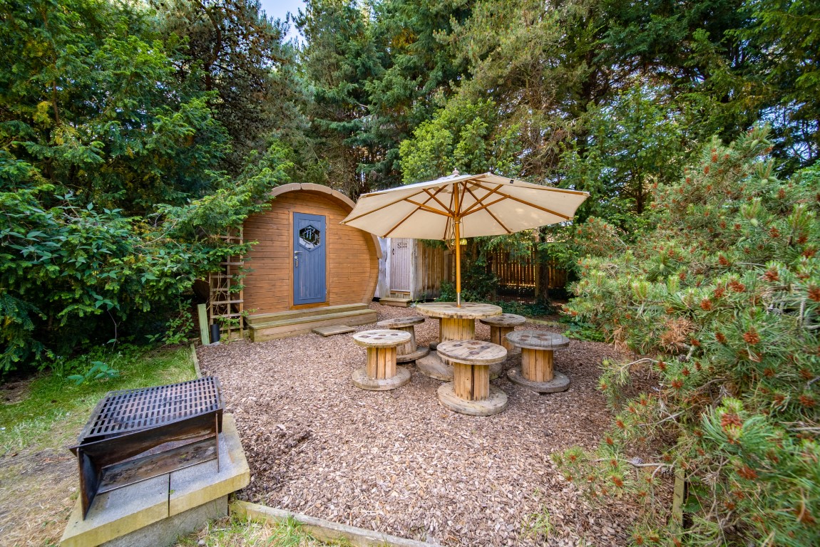 A small wooden cabin in the forested Lahtle Wood with a fenced perimeter in the background. Outside, several wooden spool tables and benches are arranged under a large patio umbrella. A grill is placed on a concrete slab nearby. Dense trees and greenery surround this perfect wood glamping spot.