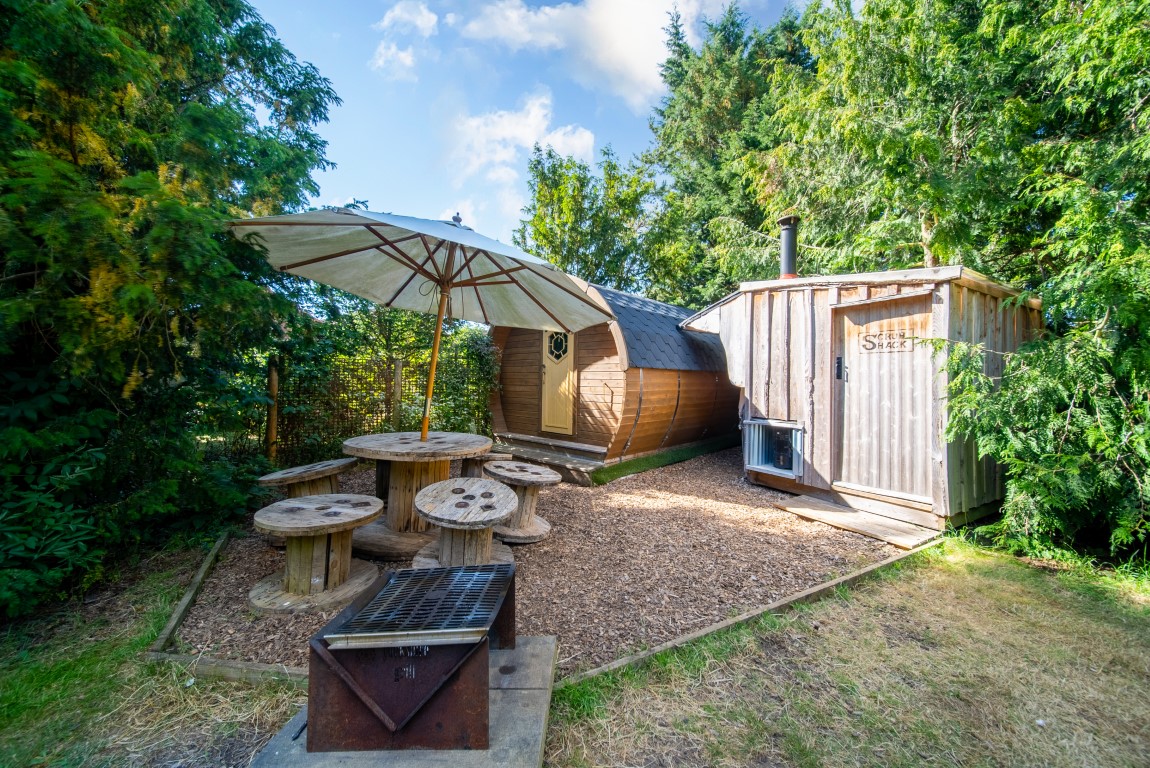 A cozy outdoor scene featuring a rustic wooden cabin surrounded by lush greenery. In front of the cabin, there's a large umbrella shading a picnic table made from wooden spools, perfect for glamping. A metal fire pit and a wooden box labeled 