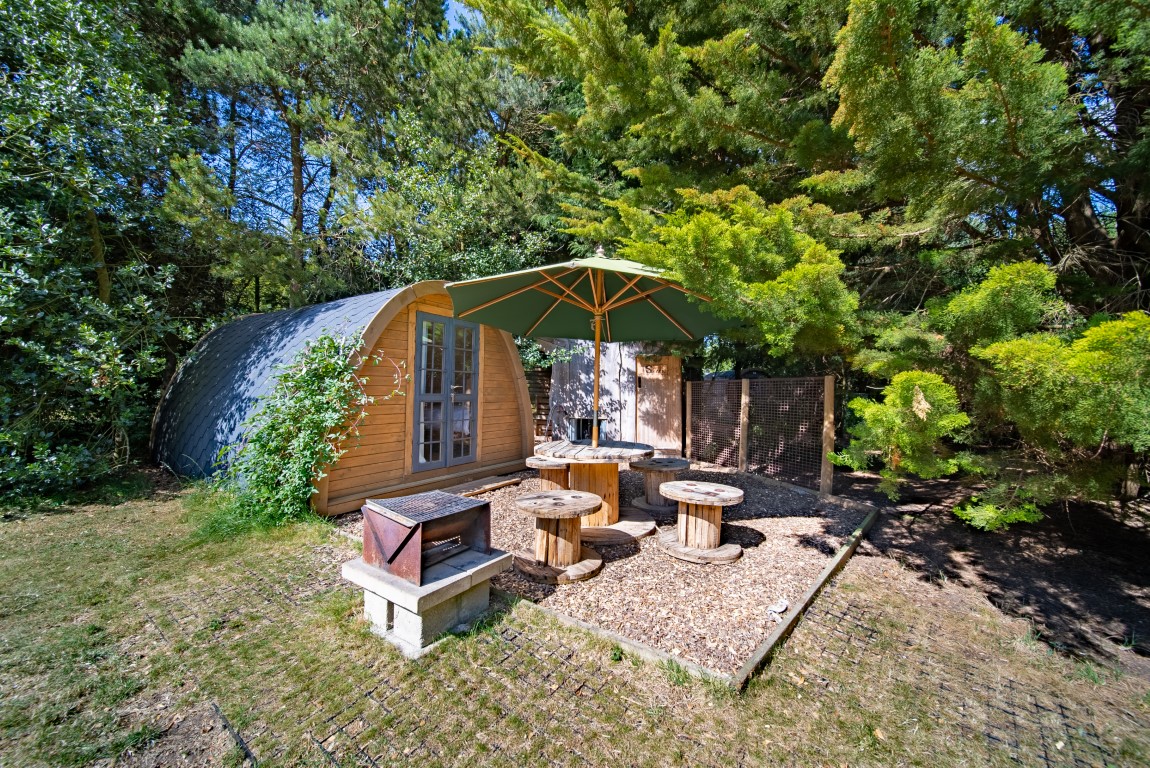 A cozy wooden cabin with a curved roof is nestled among lush green trees, perfect for glamping enthusiasts. Outside, there's a patio area with rustic wooden spool tables and seating, shaded by a large green umbrella. Nearby stands a rectangular brick barbecue on wood chip grounds—a true Lahtle escape.