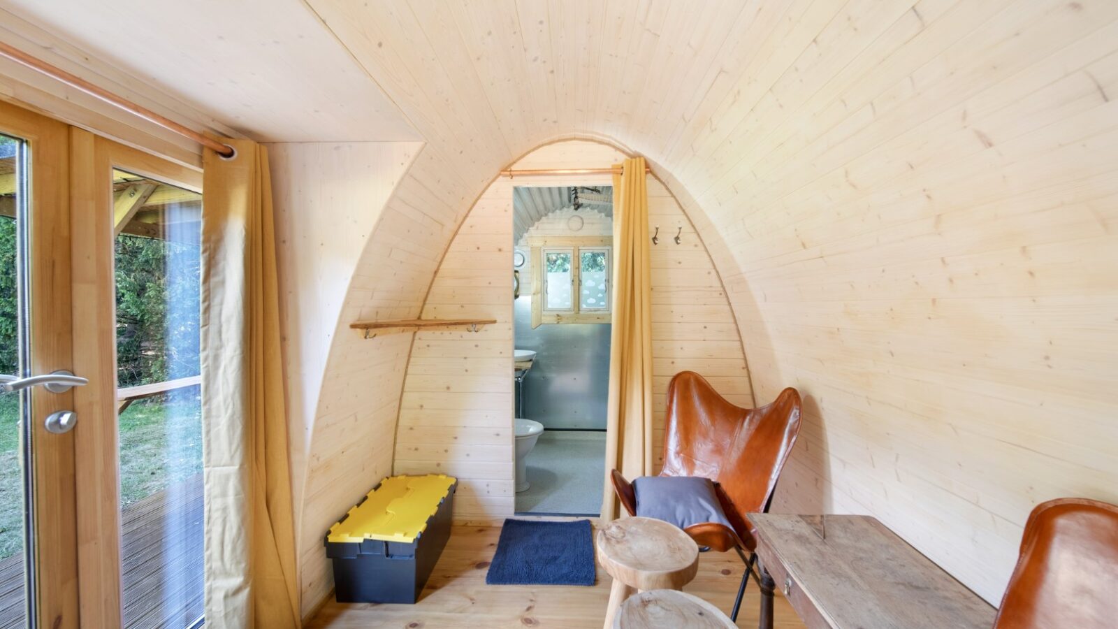 A cozy tiny house interior with light wooden walls and ceiling, perfect for glamping at Lahtle Wood. The room features a leather butterfly chair, a small wooden table, and a yellow storage bin. Large glass doors to the left let in natural light and offer a view of greenery outside. A small bathroom is visible in the background.