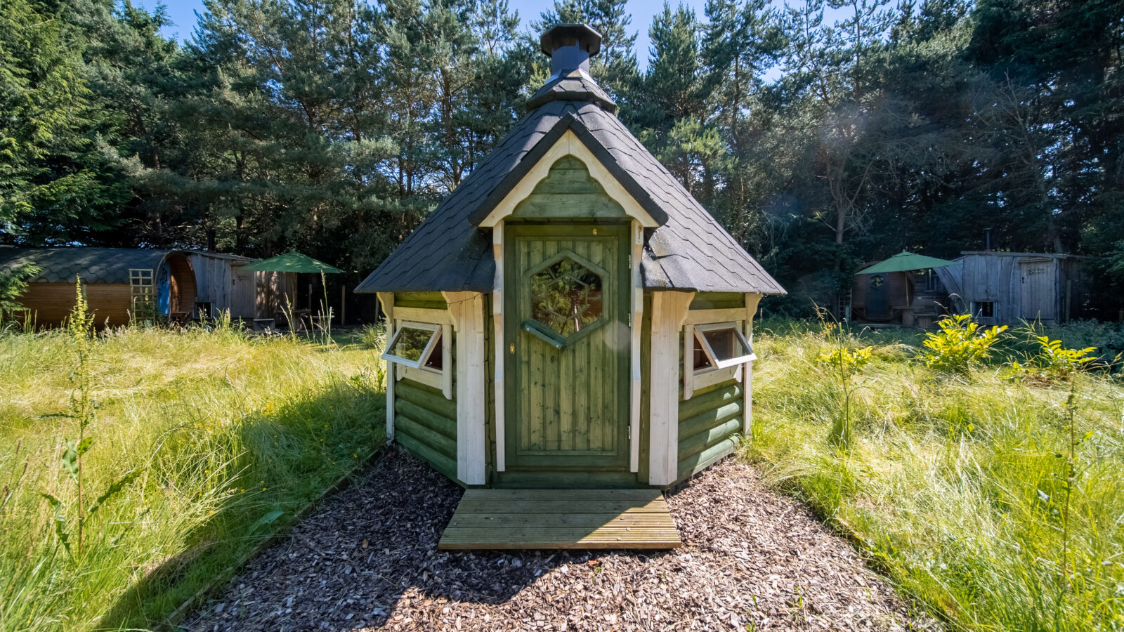 A small, wooden, green-painted cabin with a steep, shingled roof sits in a grassy clearing surrounded by pine trees in Lahtle Wood. The cabin has a front door with a decorative window, flanked by two small windows. A dirt path leads to the entrance, perfect for a serene glamping experience.