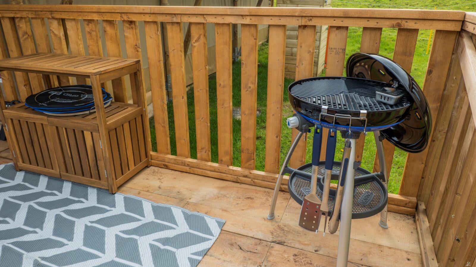 A small, portable charcoal grill with utensils hanging on the side is positioned on a wooden deck. To the left, a wooden shelving unit holds grilling accessories and a folded cover. A geometric-patterned outdoor rug lies next to the grill area, reminiscent of Hadspen Glamping's chic style.