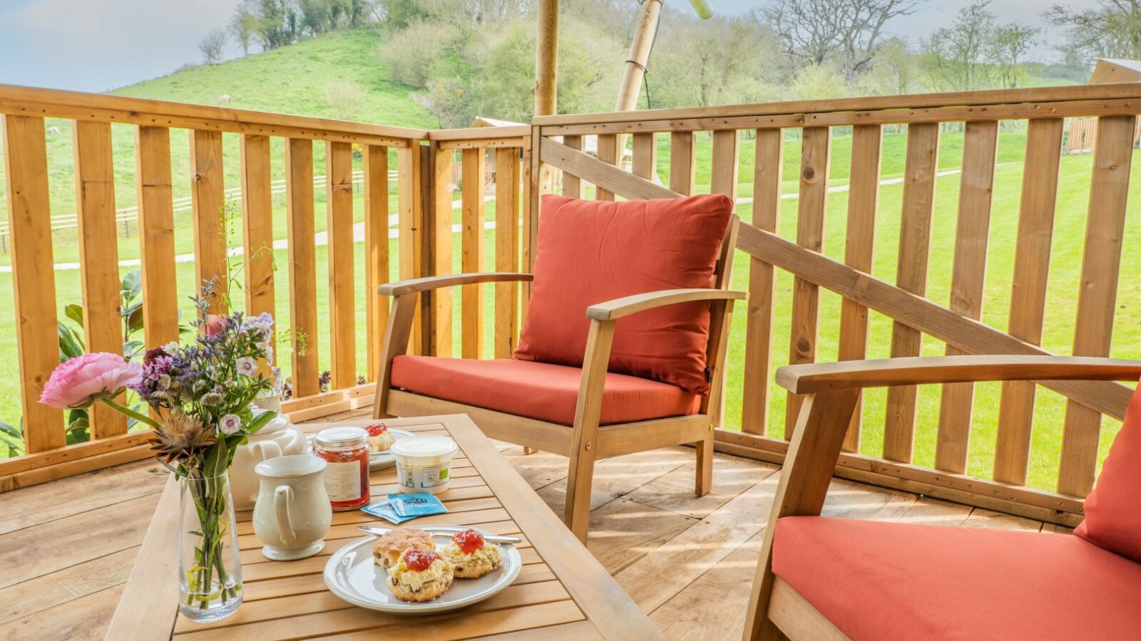 A cozy outdoor seating area with two wooden chairs featuring red cushions and a wooden table set on a wooden deck at Hadspen. The table holds a vase with flowers, a cup, jar of jam, and scones topped with cream and jam. Perfect for glamping enthusiasts, a green landscape stretches in the background.