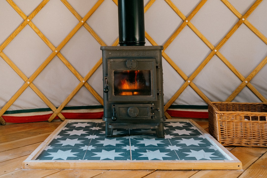 A small black wood-burning stove is set on star-patterned tiles inside the Meadow Yurt at Southleigh Farm, with wooden lattice walls surrounding it. A wicker basket is placed to the right of the stove, which has a glass window showcasing a cozy fire burning inside.