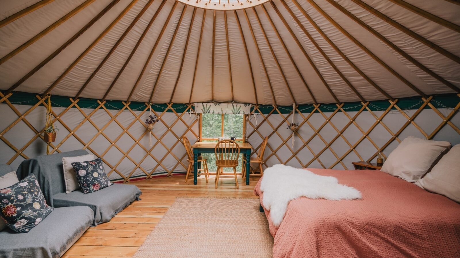 Interior of the Meadow Yurt at Southleigh Farm featuring wooden lattice walls, a skylight, and wood flooring. The space includes a bed with pink bedding, a small table and chair by the window, and seating with floral cushions, creating a warm and inviting atmosphere.