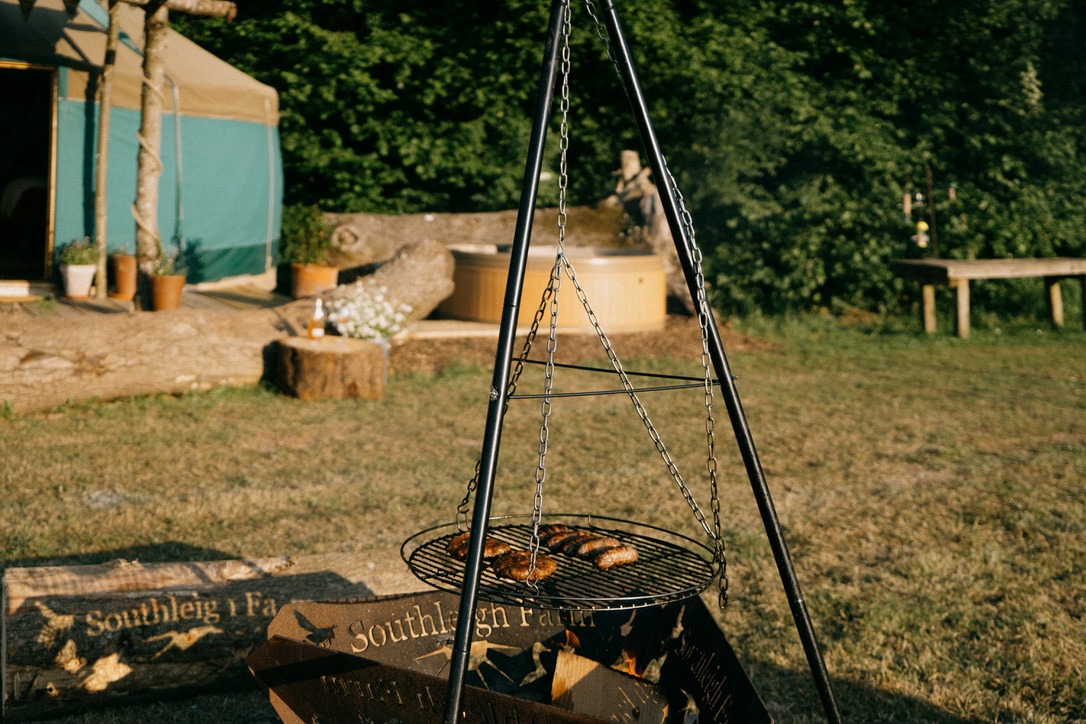 A triangular metal grill stand suspended over a fire pit with food cooking on the grill. Behind it, at Southleigh Farm, there's a canvas tent, wooden logs, and outdoor seating with greenery in the background, creating a rustic camping scene.